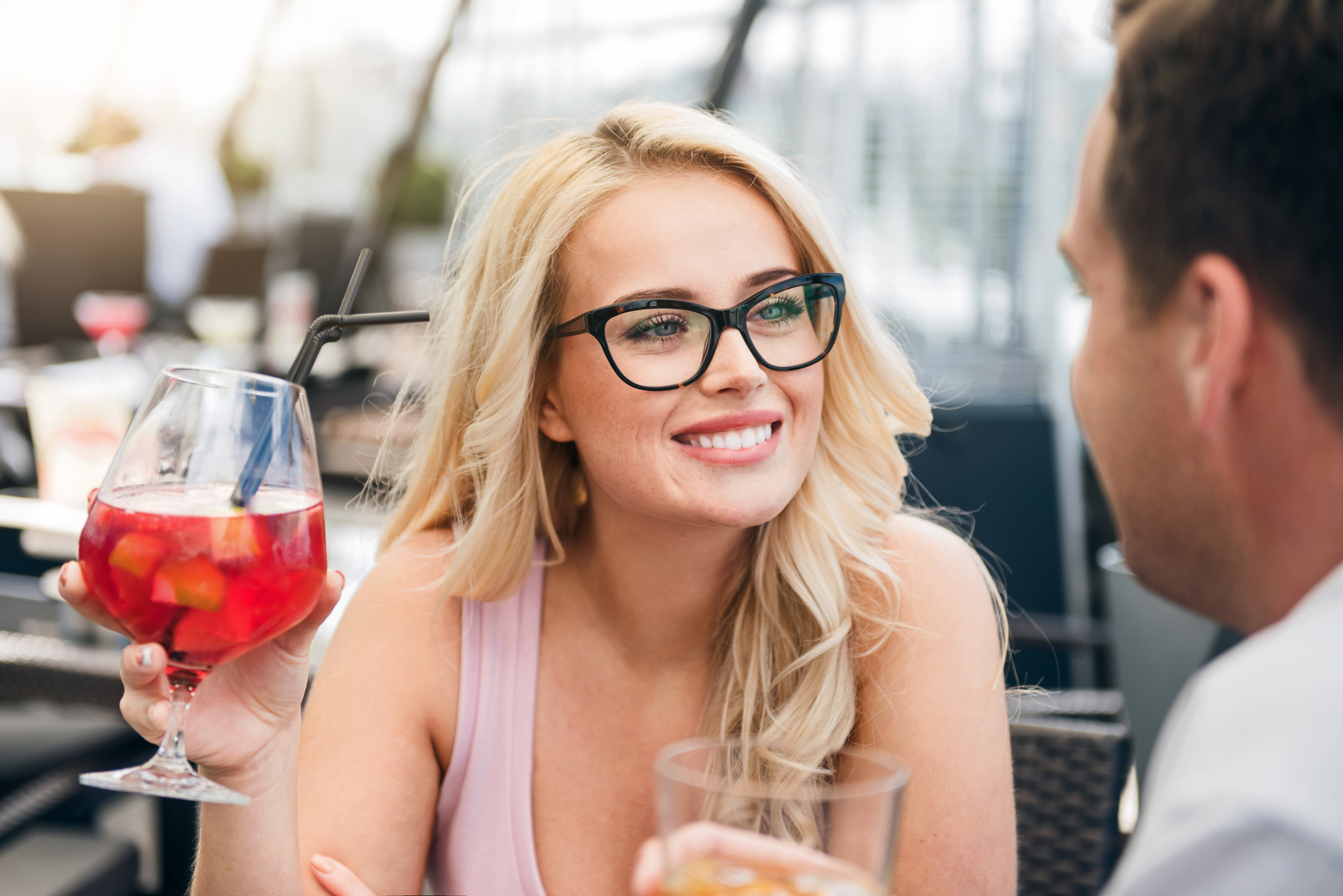 A woman with long blonde hair and glasses is holding a glass of red sangria with fruit pieces. She is smiling and looking at a man sitting across from her. They appear to be at an outdoor restaurant or cafe.