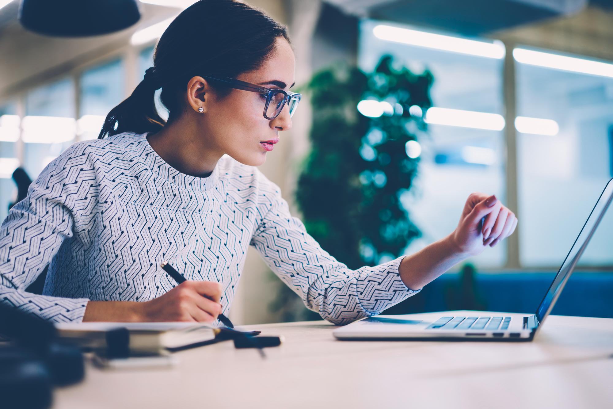 A focused woman in a white patterned sweater and glasses is sitting at a desk in an office. She is working on a laptop, taking notes with a pen, and appears to be deeply engrossed in her task. The background shows an office setting with windows and plants.