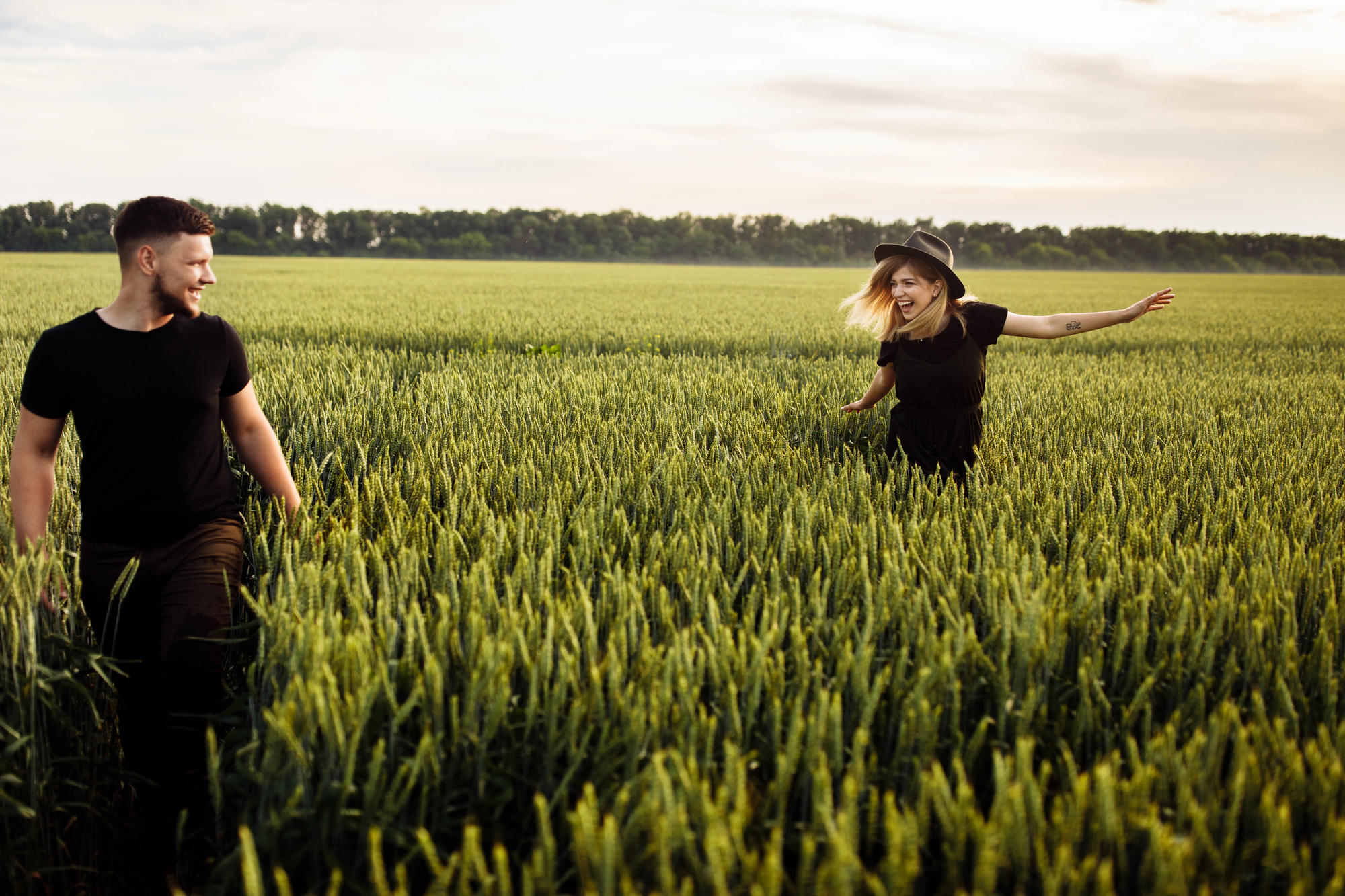 A man and woman, both wearing black clothing, traverse through a lush green field of tall wheat. The woman, wearing a hat, has her arms outstretched and smiles at the man, who looks back at her. A line of trees is visible in the background under a golden sky.