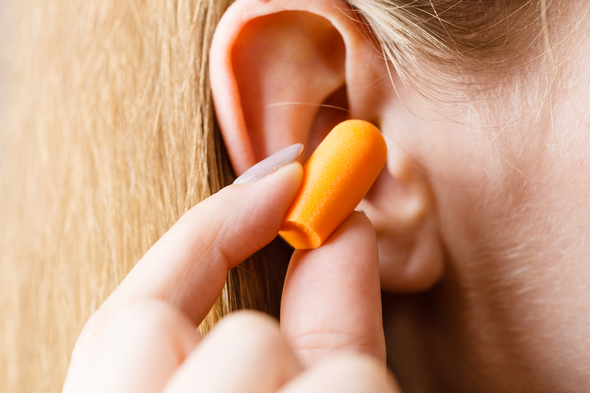 A close-up image of a person inserting an orange earplug into their ear. The earplug is being held between the thumb and index finger. The person has light-colored hair.