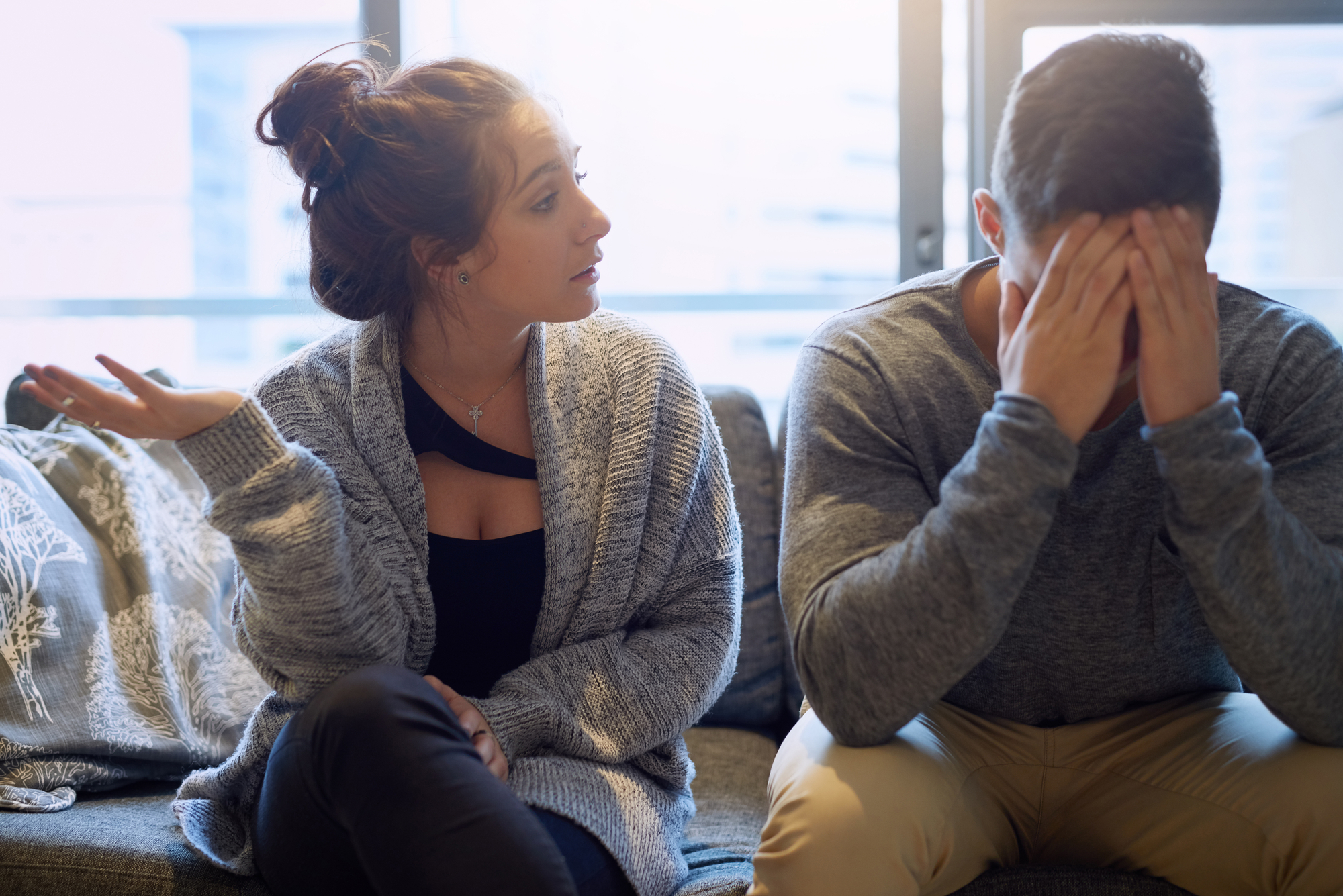 A woman gestures with frustration while sitting on a couch next to a man who is covering his face with his hands. The man appears to be distressed, and they are in a room with a large window in the background.