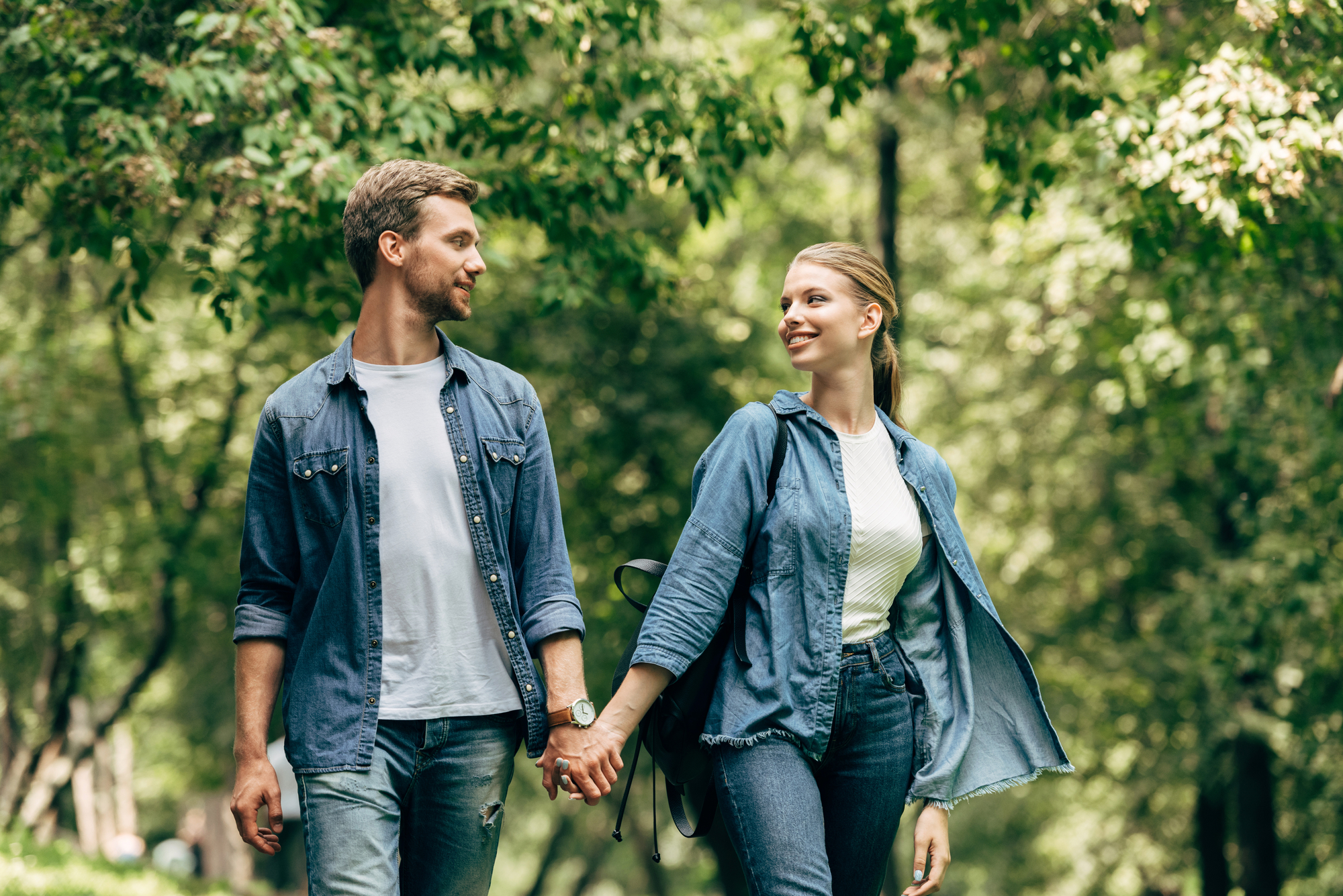 A couple holds hands while walking outdoors in a green, leafy park. Both are casually dressed in denim jackets and jeans. They are looking at each other and smiling, enjoying a sunny day together.