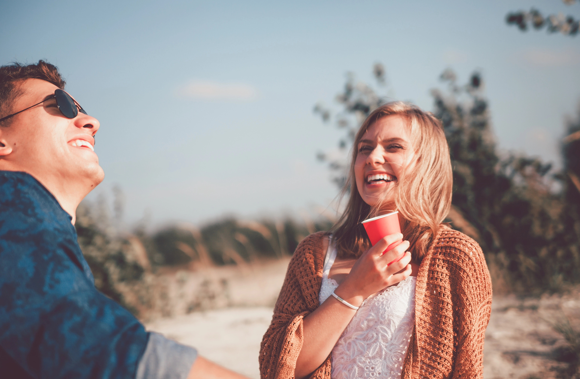 A man and woman enjoy a sunny day outdoors. The man is wearing sunglasses and smiling, while the woman, dressed in a light brown cardigan over a white top, holds a red cup and laughs. The background shows greenery and a clear blue sky.