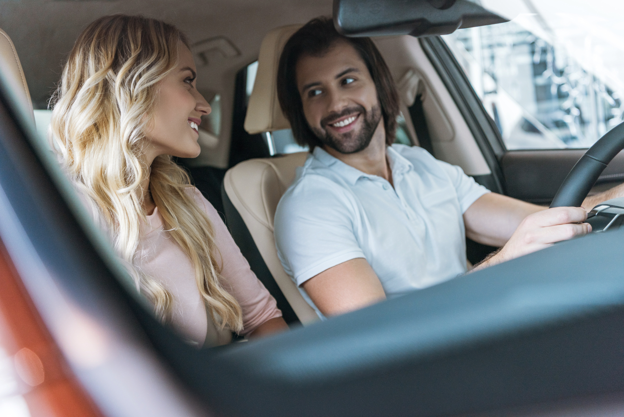 A man and a woman sit in a car, smiling at each other. The man, wearing a white shirt, is in the driver's seat with his hands on the steering wheel. The woman, with long blonde hair and a pink top, is in the passenger seat looking at him. Both appear happy and relaxed.