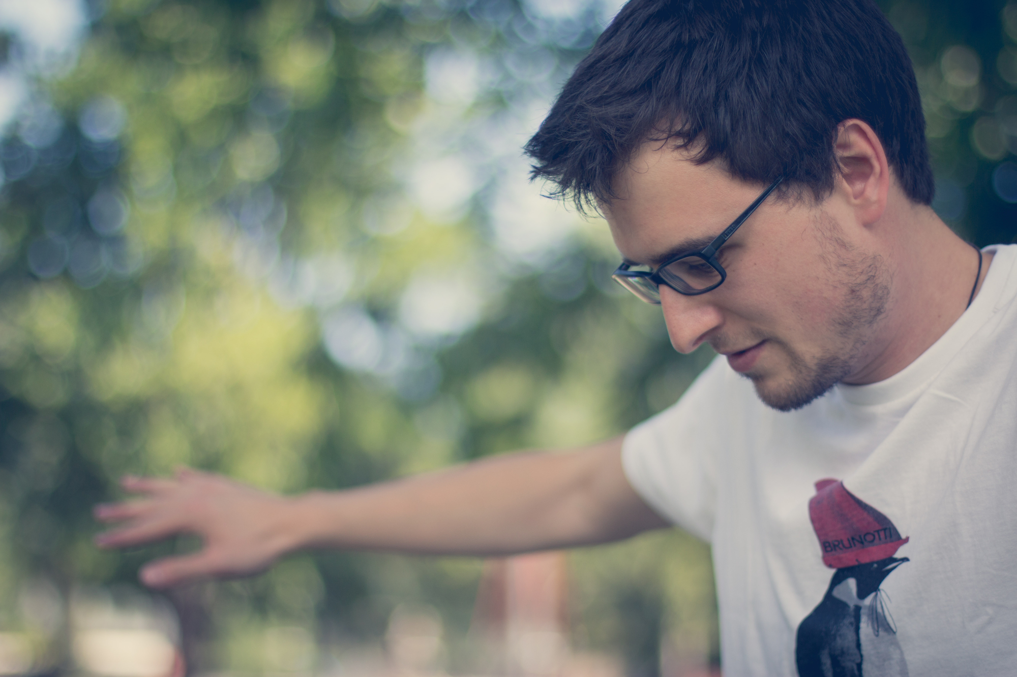 A young man with short dark hair and glasses stretches his arms out to the sides. He is wearing a white t-shirt with a graphic design. The background is blurred, featuring green foliage from trees. The image has a soft, serene ambiance.