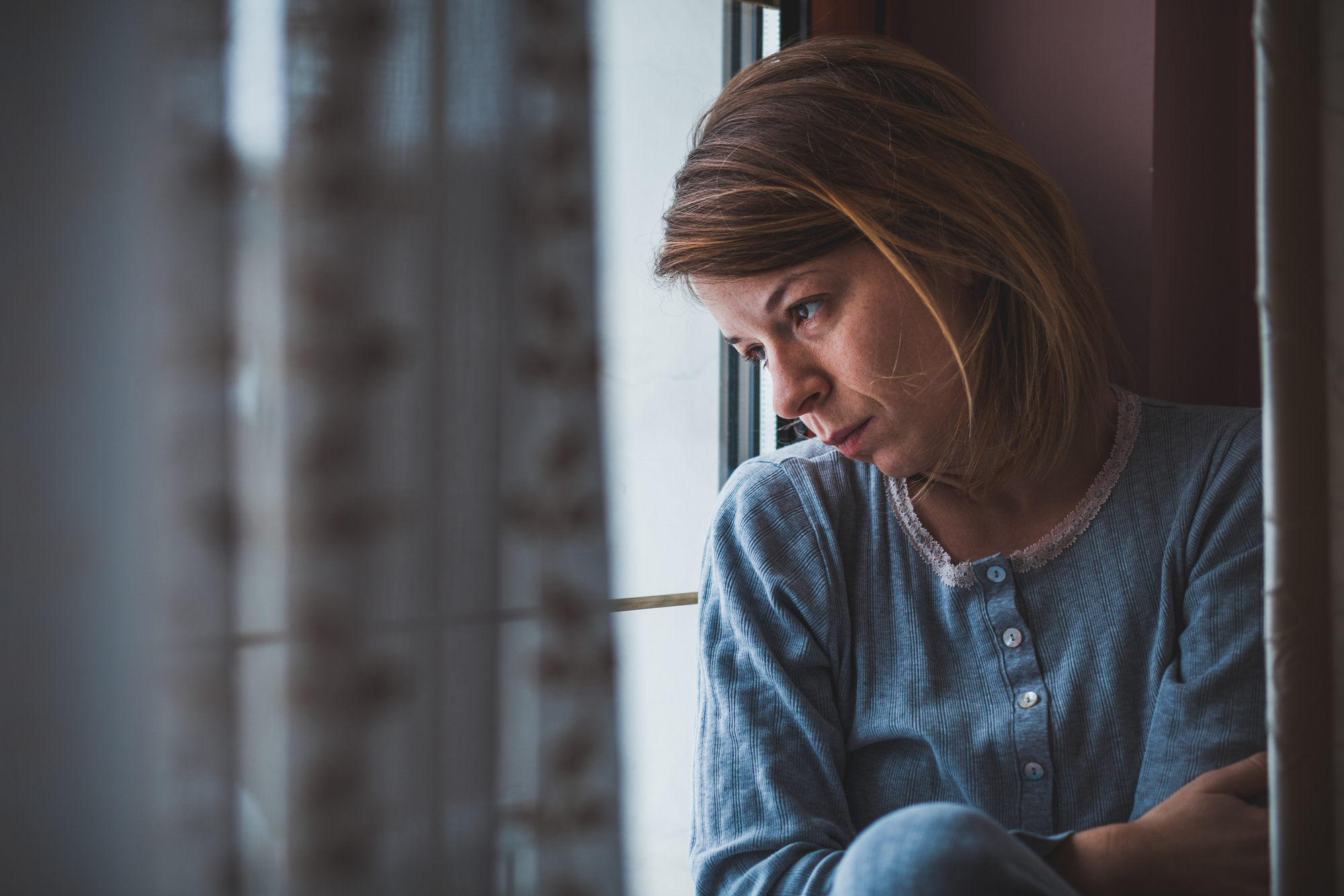 A woman wearing a blue top sits by a window with a distant, contemplative expression. Her arms are crossed over her knees, and light filters through lace curtains nearby, casting a soft glow on her face. The scene evokes a mood of introspection and melancholy.