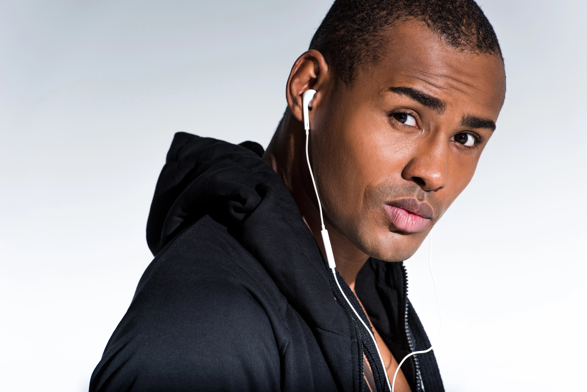 A person wearing a black hoodie is listening to music through white earphones. They have a serious expression on their face and are looking slightly off to the side. The background is a plain gradient from white to light gray.