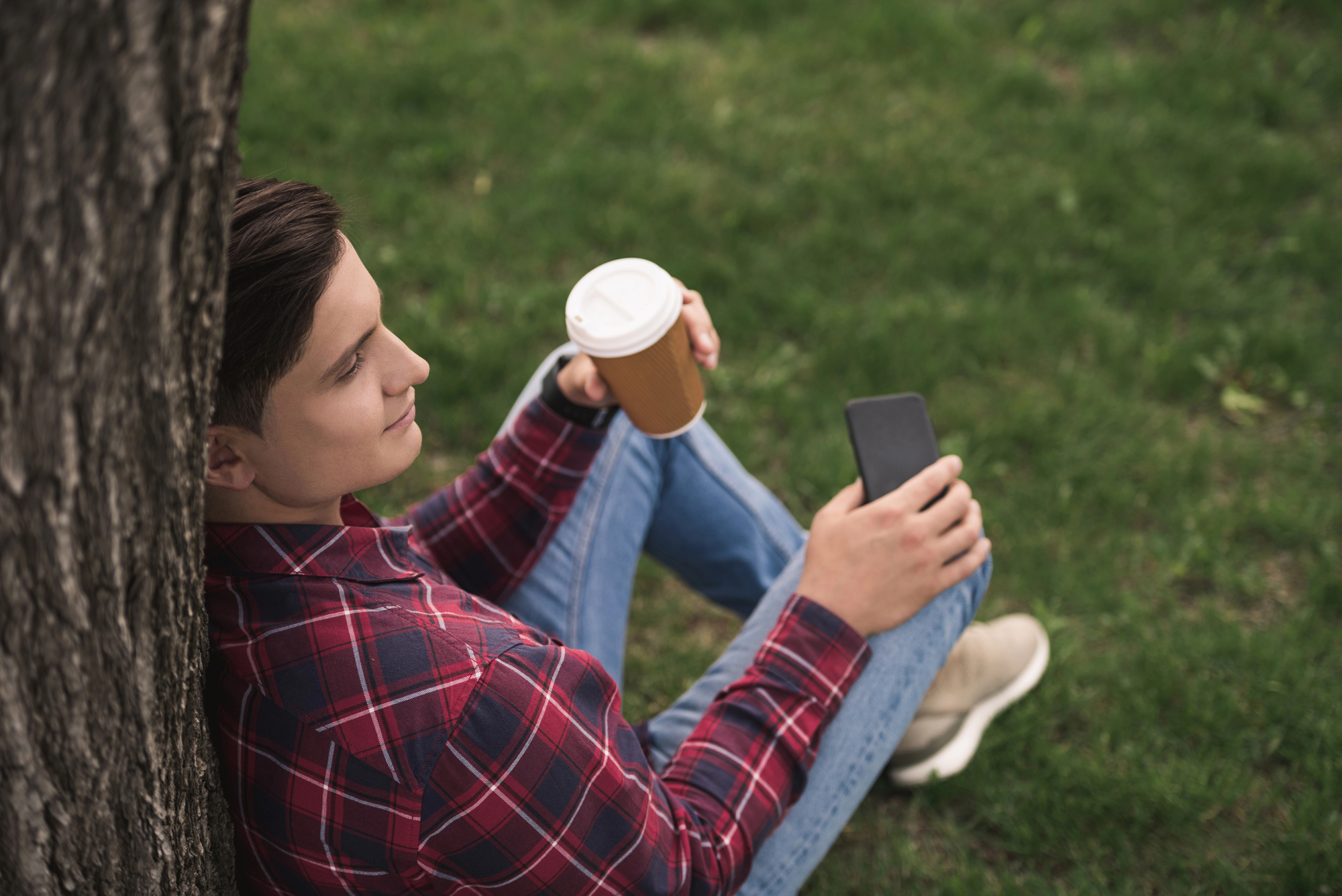 A person wearing a red plaid shirt and blue jeans sits on grass, leaning against a tree. They are holding a to-go coffee cup in one hand and a smartphone in the other, looking content and relaxed. The background is grassy, indicating a park or outdoor space.