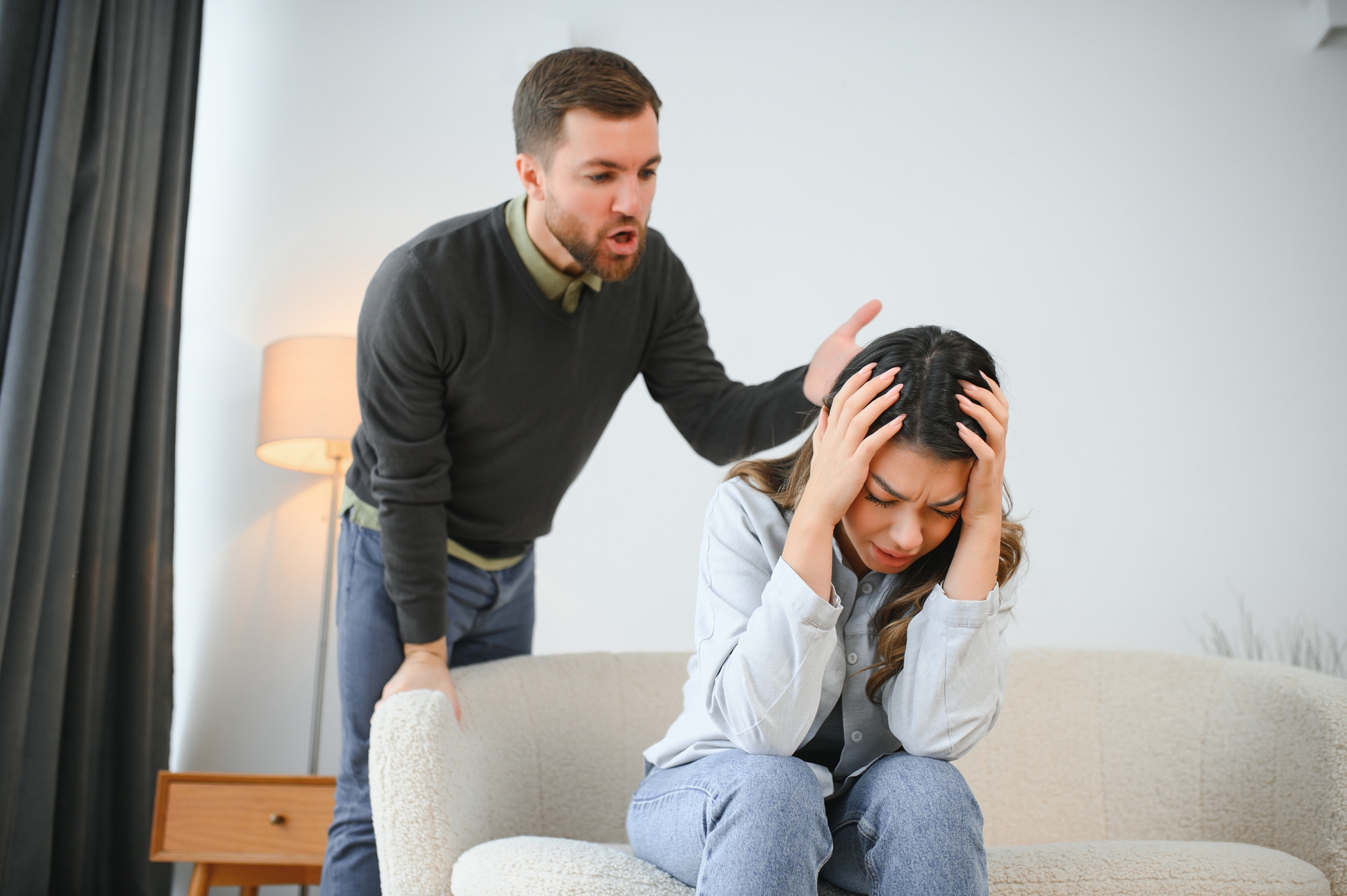 A man with a beard is shouting at a woman sitting on a sofa. The woman, distressed, is holding her head in her hands. The setting is a modern living room with a lamp and side table in the background.