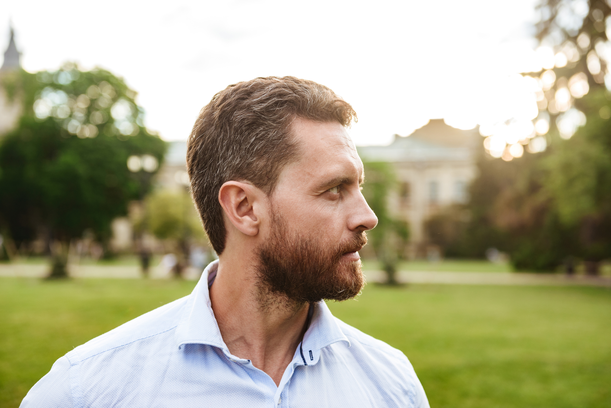 A man with short, curly hair and a beard, wearing a light blue shirt, gazes to his left. He is standing outdoors in a lush green park with trees and buildings in the background, under a partly cloudy sky.