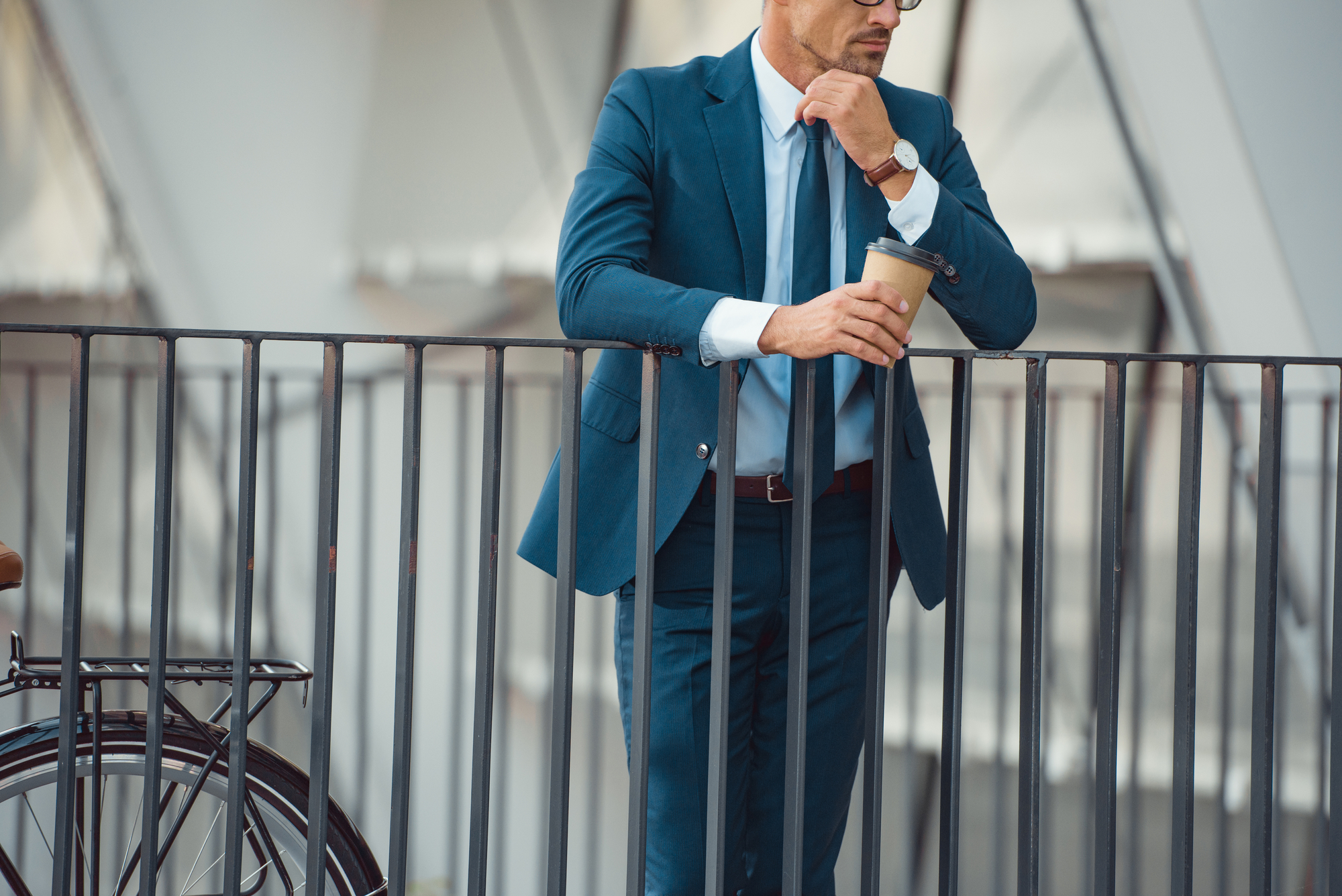 A man wearing a blue suit and tie leans on a metal railing, holding a coffee cup and appearing deep in thought. A bicycle is partially visible to the left. The setting appears to be an urban environment, possibly near office buildings.