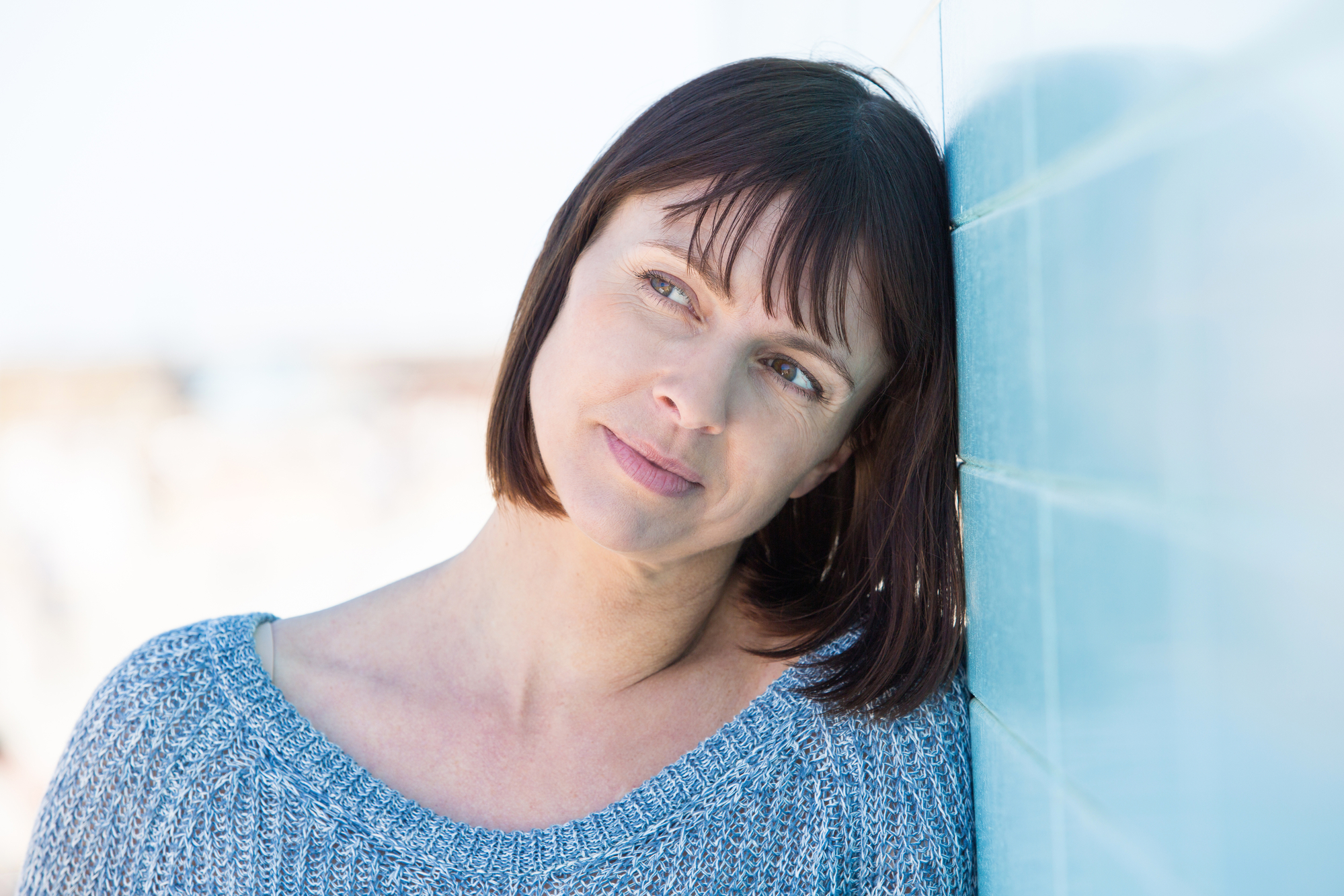 A woman with short brown hair, wearing a light blue knit sweater, leans her head against a blue tiled wall. She gazes off to the side with a thoughtful expression. The background is blurred and suggests an outdoor setting.