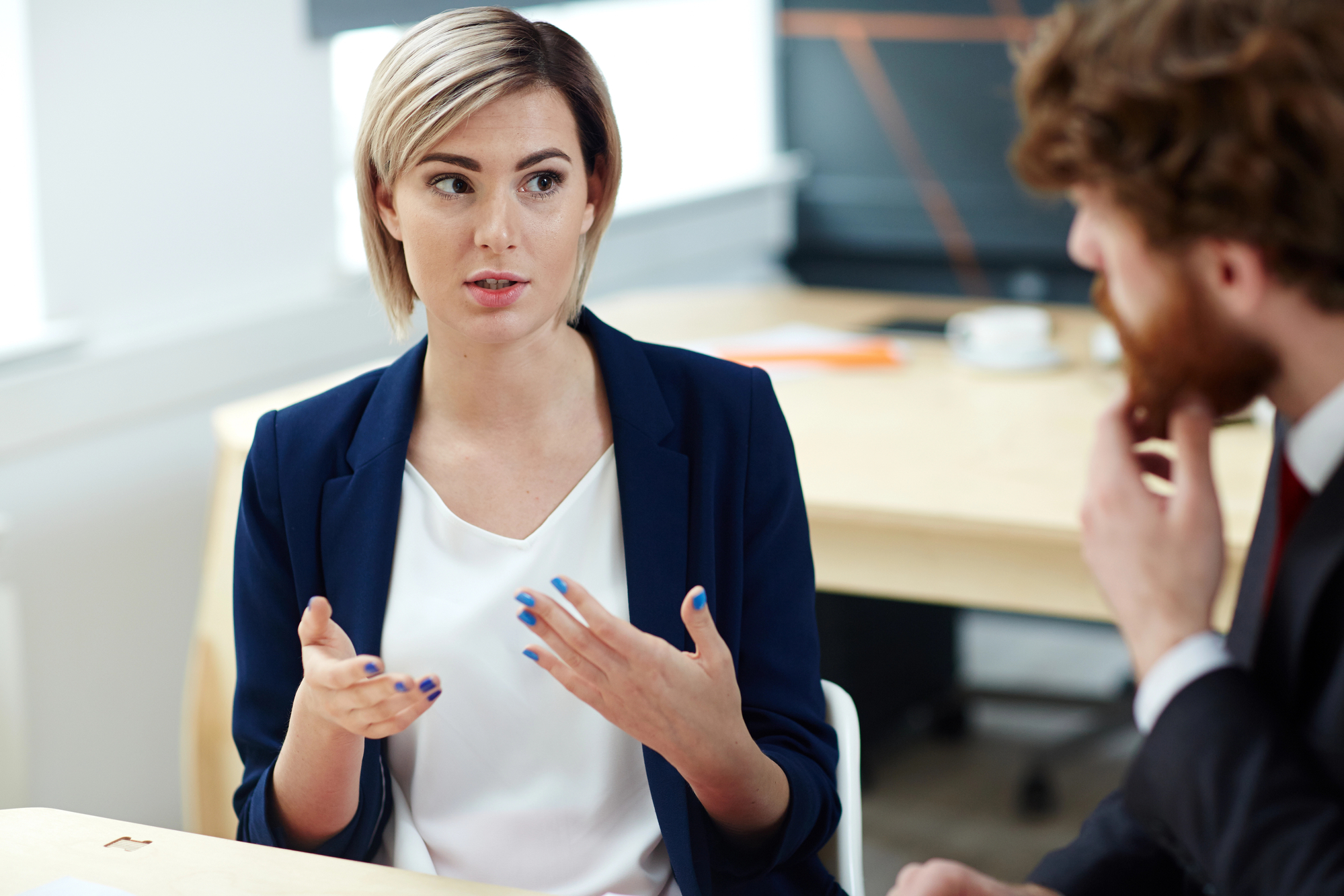 A woman with short blonde hair, wearing a navy blazer and white top, sits at a desk and gestures while talking to a man with curly hair and a beard who is listening intently. They are in a bright office setting with windows in the background.