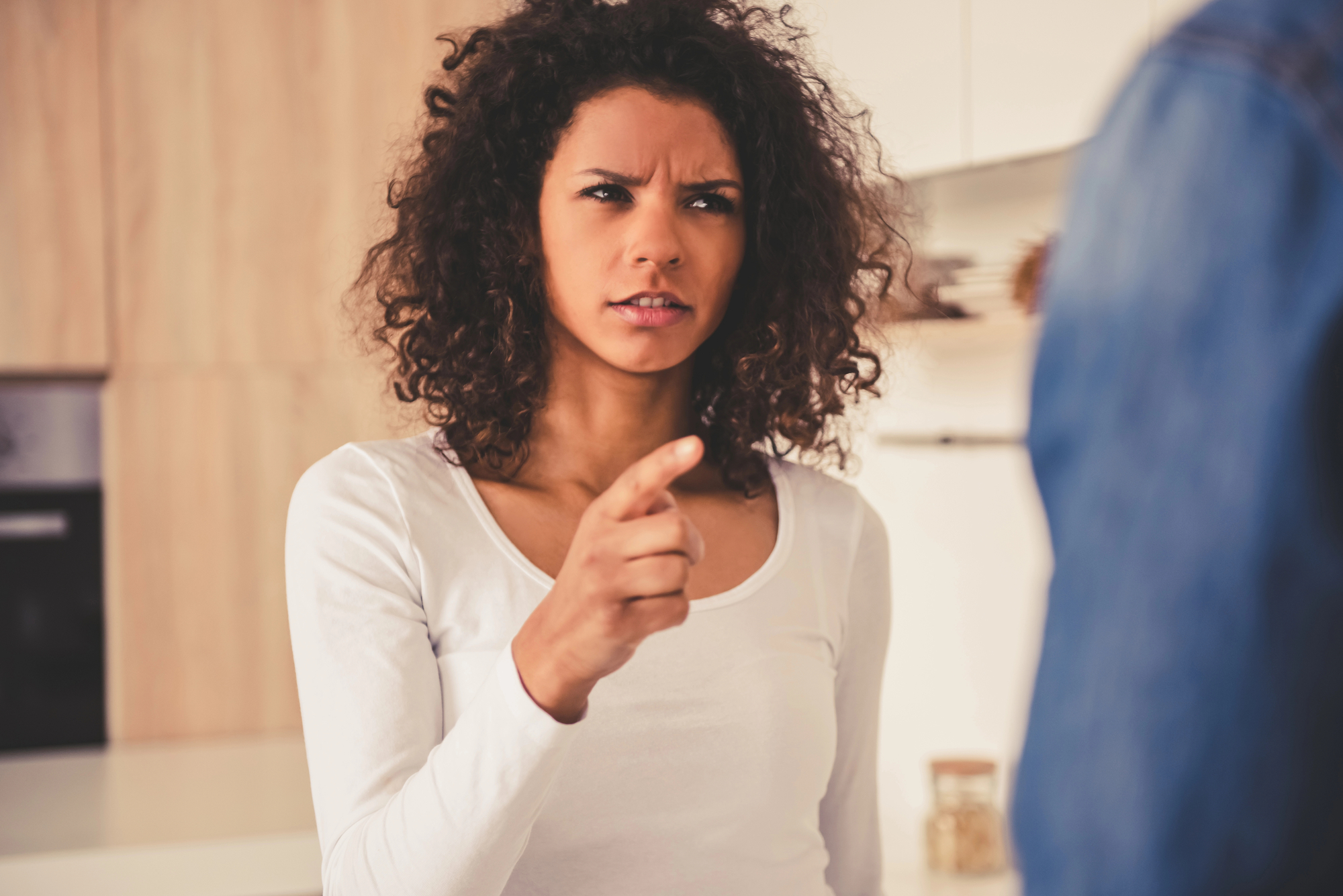 A woman with curly hair, wearing a white long-sleeved shirt, points her index finger at someone out of the frame. She has an intense and serious expression on her face. The background appears to be a bright, modern kitchen.