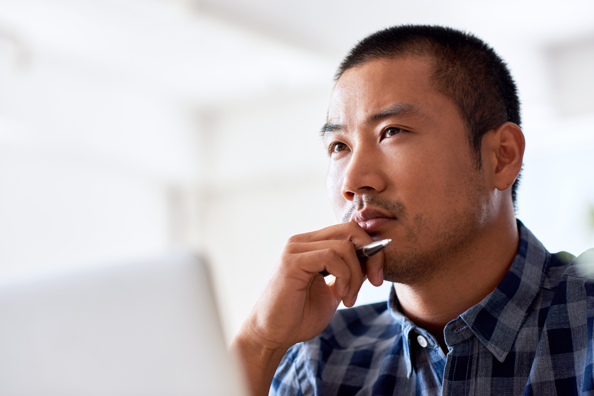 A focused man with short black hair sits in a bright room, resting his chin on his hand while holding a pen. He is wearing a blue and white checkered shirt and is looking thoughtfully into the distance, possibly contemplating something on his laptop screen.