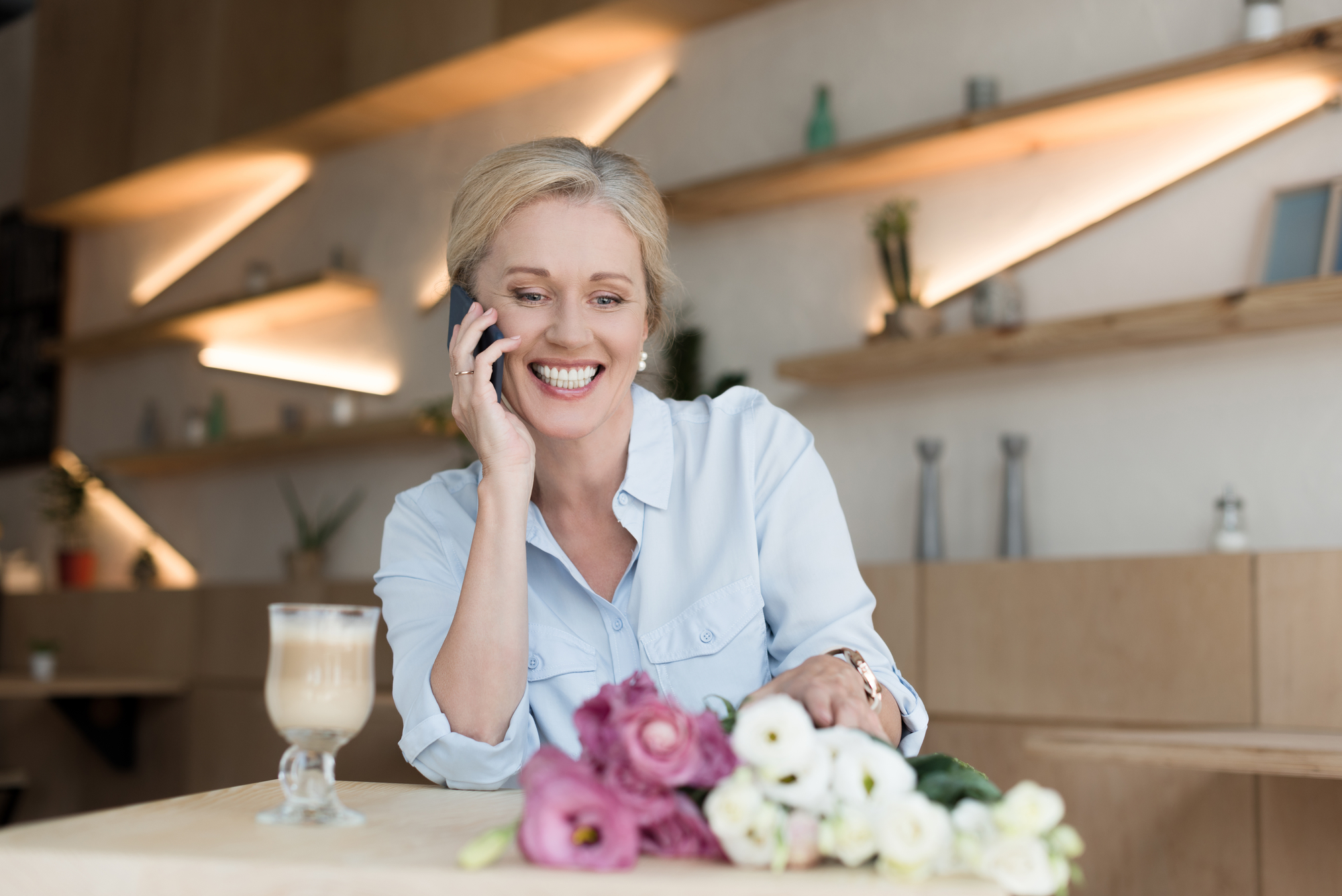 A woman with blonde hair smiles while talking on her cellphone in a modern, warmly lit cafe. She wears a light blue shirt and touches a bouquet of pink and white flowers on a table. A latte sits in front of her. The background features geometric shelves with decor.