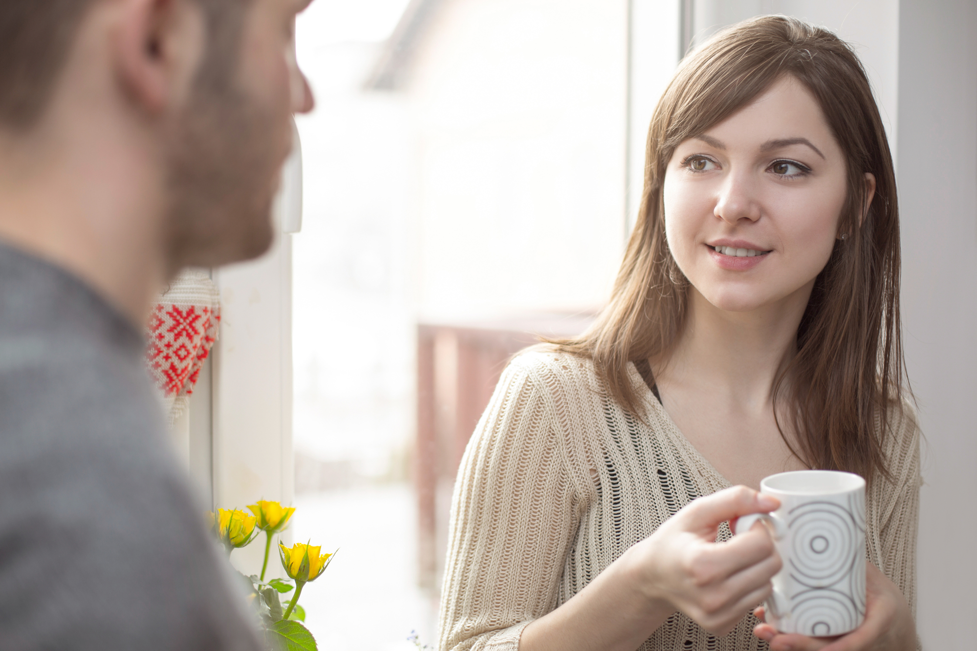 A woman with long brown hair, holding a white mug with circular patterns, smiles and talks to a man with a beard. They are standing by a window with yellow flowers on the windowsill, and an embroidered cloth is partially visible in the background.