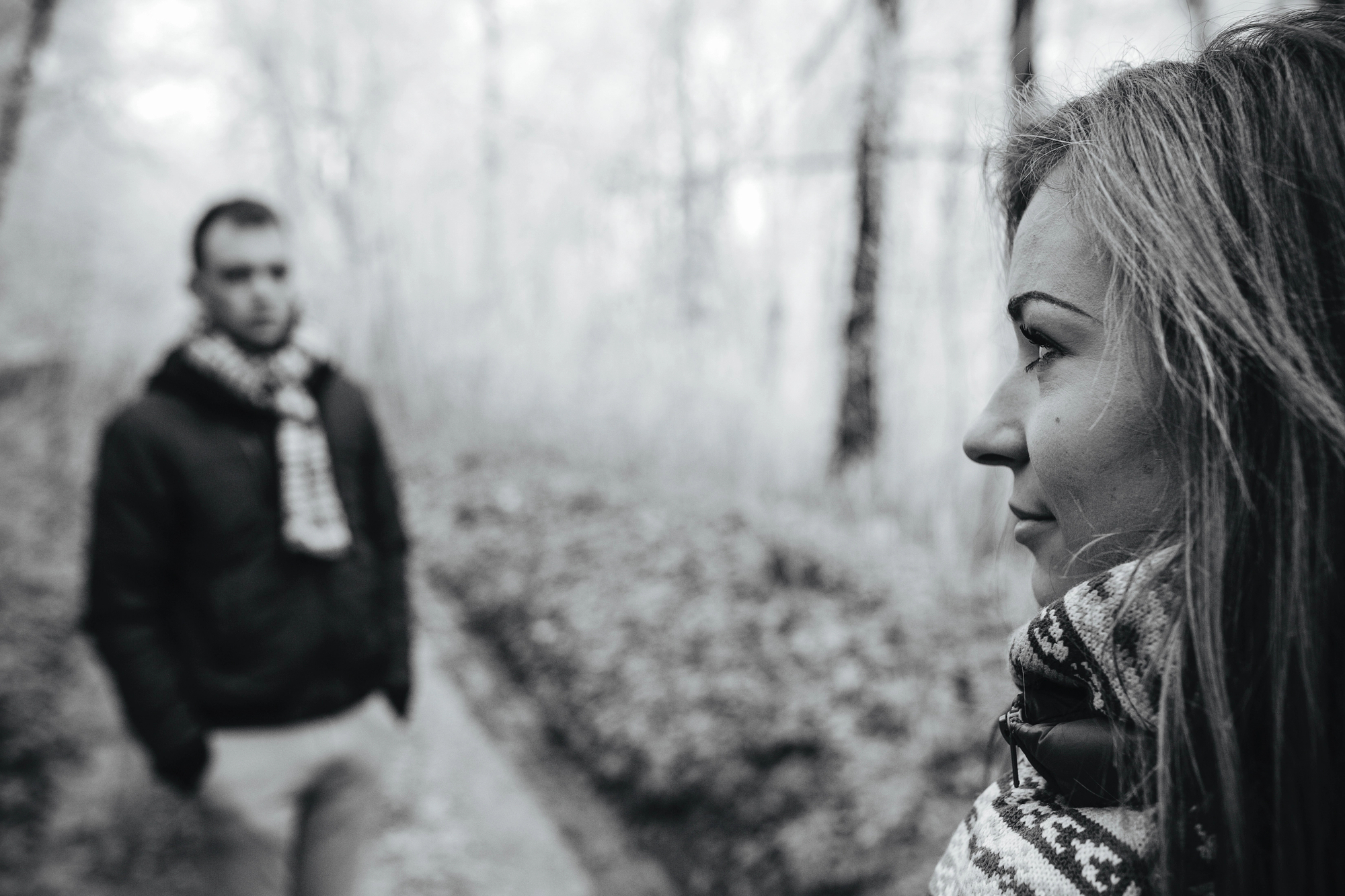 A black and white photo of a woman in the foreground looking to the side with a pensive expression. In the blurred background, a man stands on a path surrounded by trees. Both are wearing scarves, suggesting a cool day.