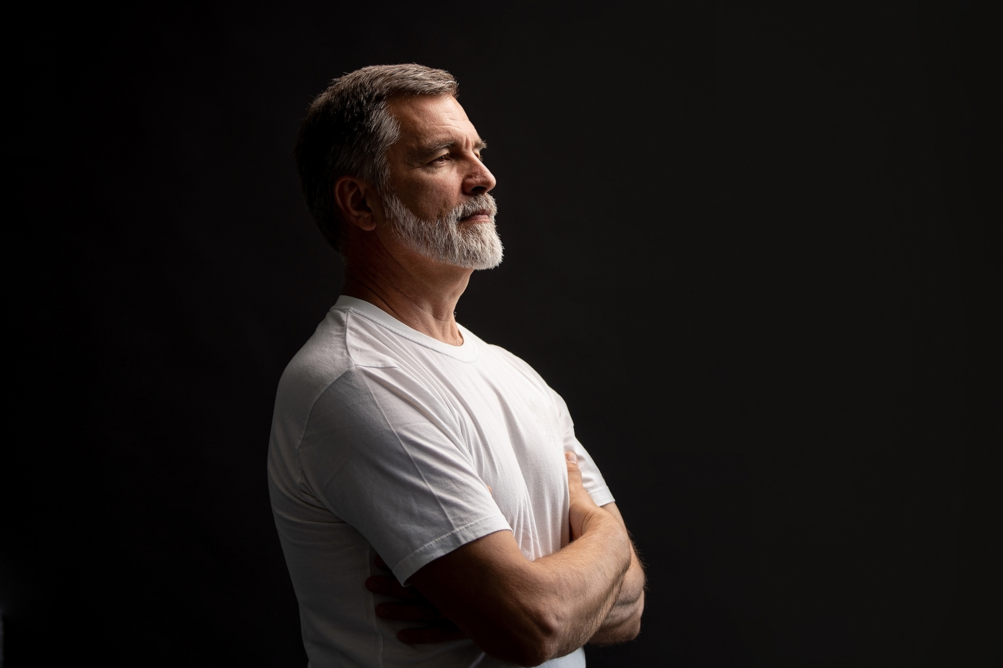 A man with gray hair and a beard is standing with his arms crossed, facing to the side against a dark background. He is wearing a white t-shirt and appears thoughtful.
