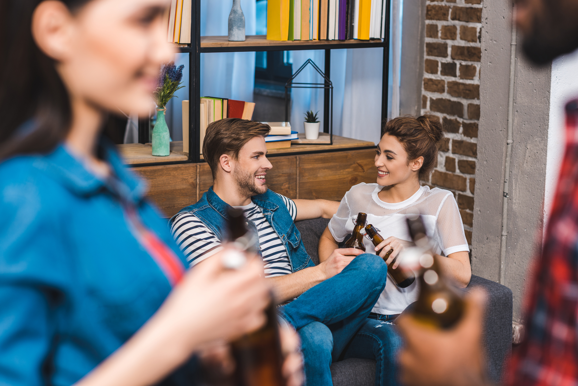 A group of friends enjoying time together indoors. A man and a woman are sitting on a couch, smiling and holding beer bottles. Two other people are standing in the foreground, each holding bottles. A bookshelf with colorful books and decorations is in the background.