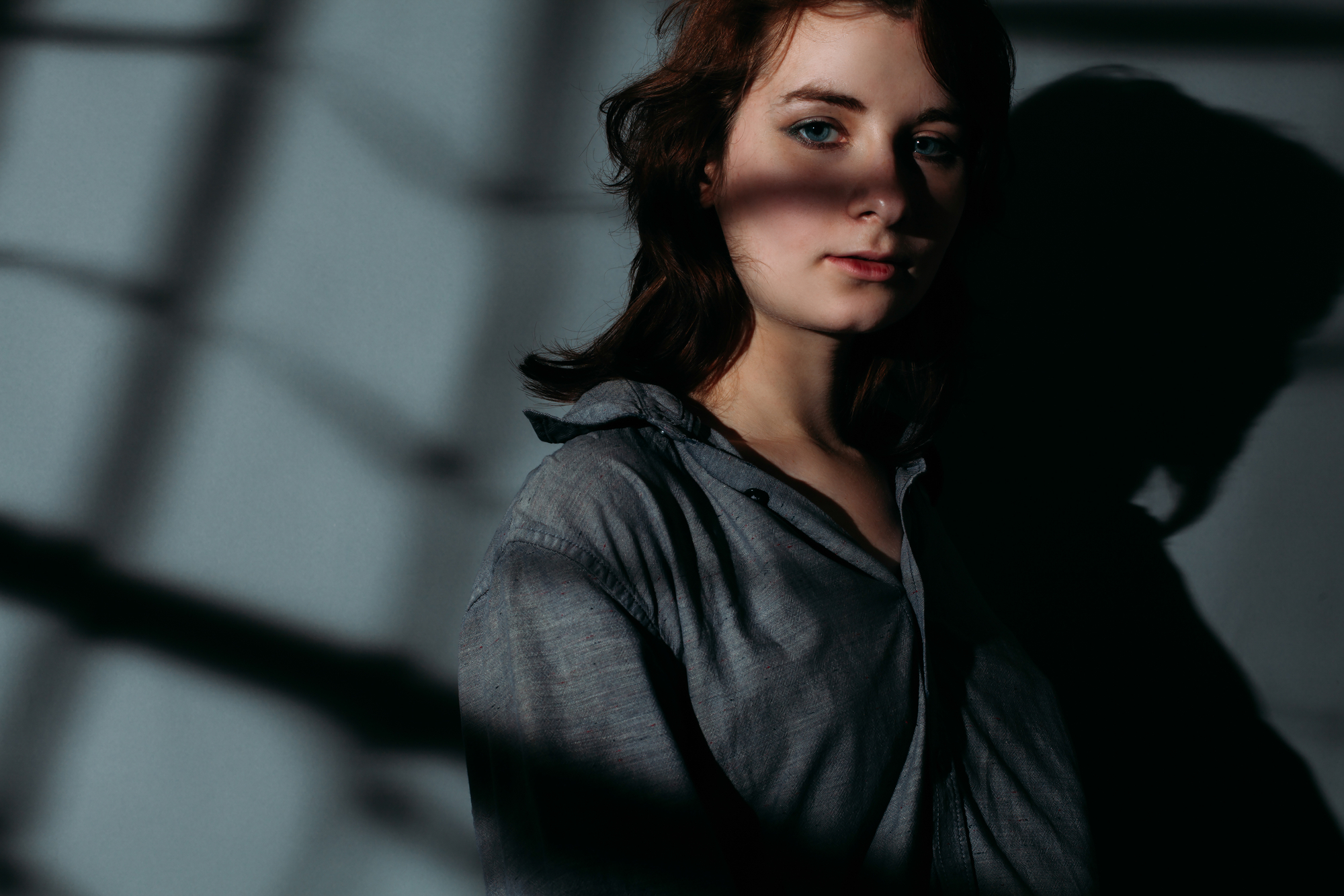 A person with shoulder-length hair stands against a wall with shadows cast over them. They are wearing a loose, button-up shirt and looking pensively at the camera. The lighting creates a dramatic contrast with the background.