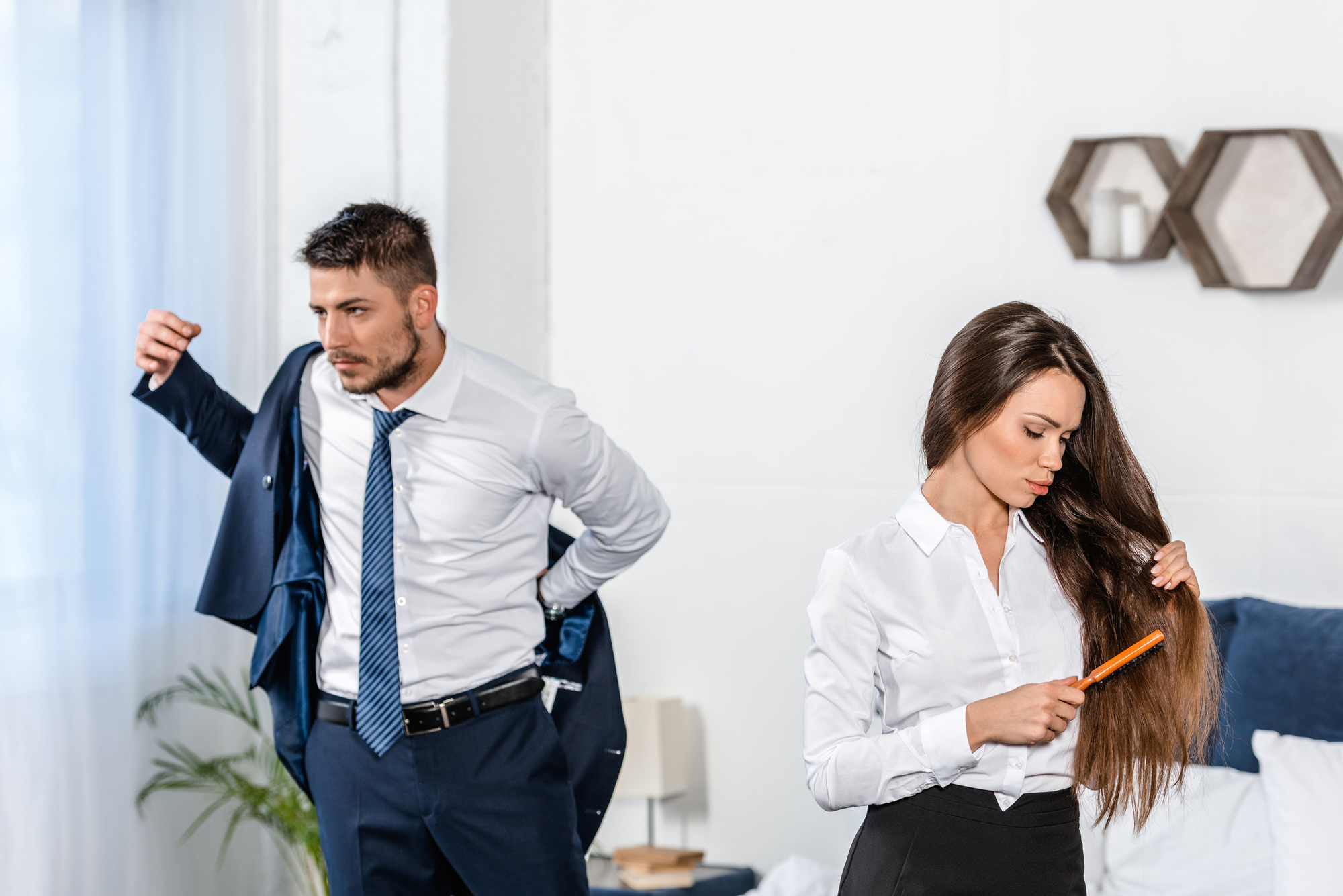 A man is putting on a jacket while a woman combs her long hair. Both are wearing business attire, with the man in a suit and tie and the woman in a white blouse and black skirt. They appear to be getting ready, standing in a well-lit, modern room.