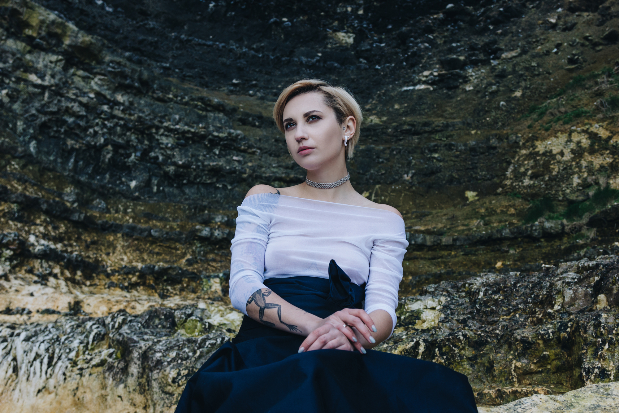 A person with short blonde hair sits against a rocky background, wearing a white off-the-shoulder top and a black skirt. They have a tattoo on their left arm and are looking slightly off to the side with a contemplative expression.