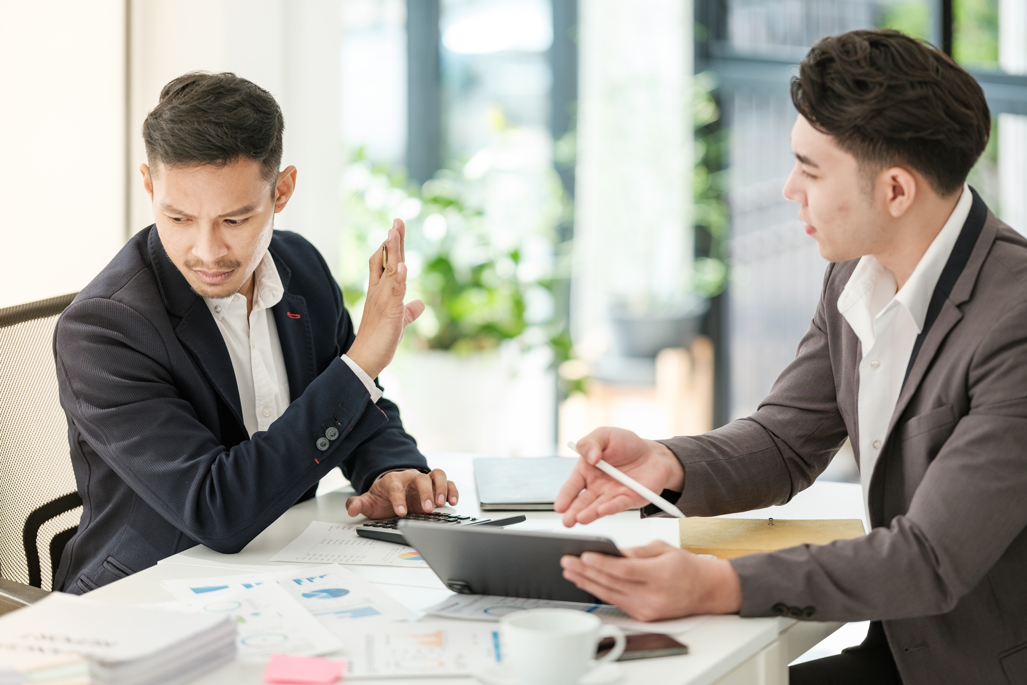 Two men in business attire sit at a desk cluttered with paper and documents. The man on the left gestures with a raised hand, appearing to dismiss or reject something. The man on the right holds a pen and tablet, engaging in a discussion.
