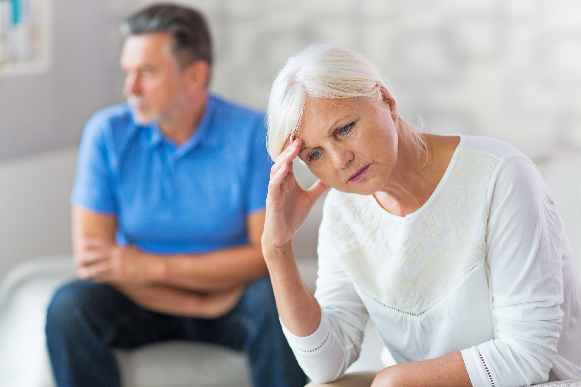 A senior woman with white hair in a white shirt appears contemplative, holding her head with one hand. A senior man with gray hair in a blue shirt sits in the background, looking away. Both appear to be deep in thought or having a disagreement.