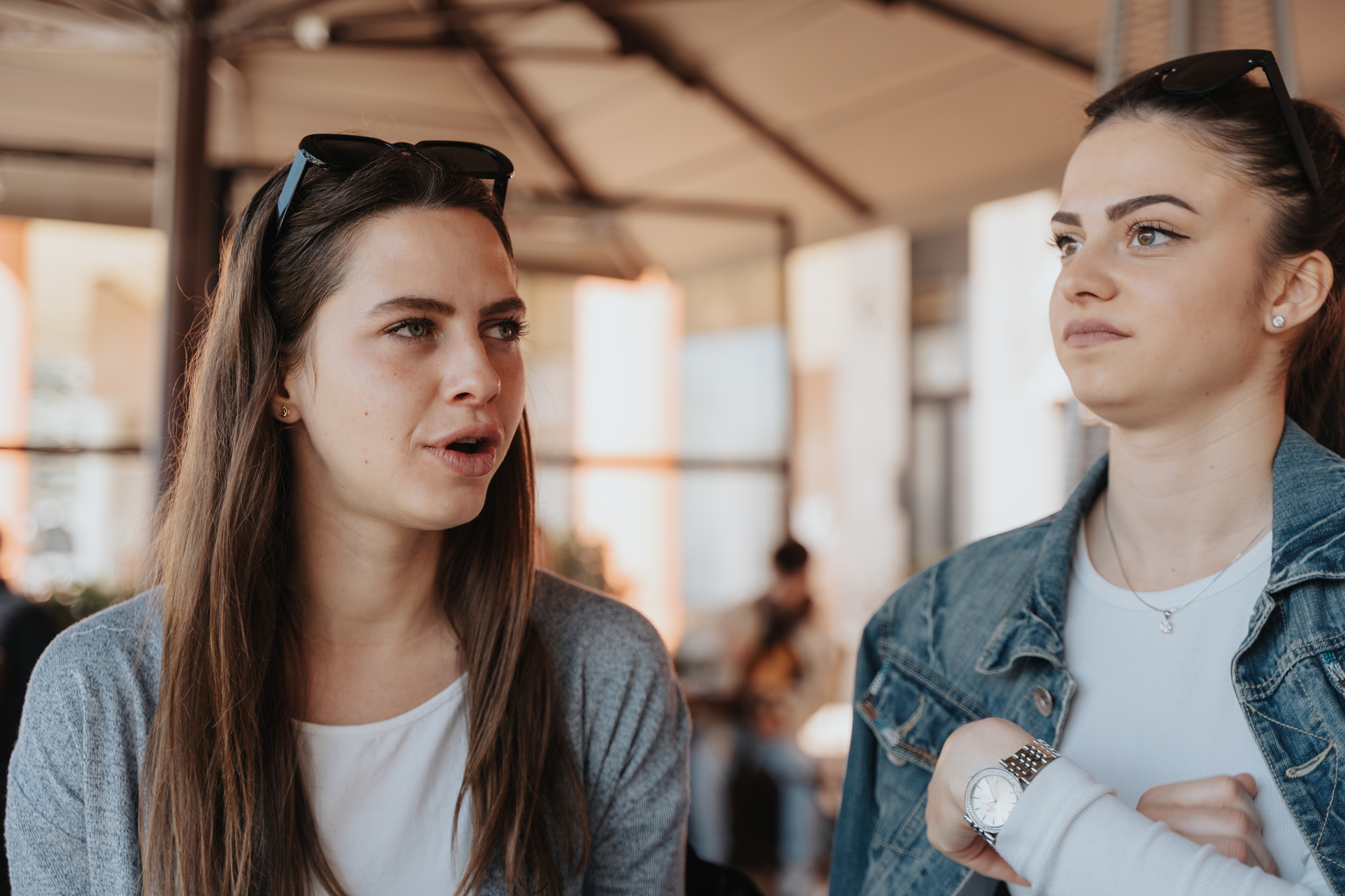 Two women are seated and engaged in conversation at an outdoor café. The woman on the left is speaking, while the woman on the right listens attentively with a thoughtful expression. Both have long hair and are casually dressed, with sunglasses resting on their heads.