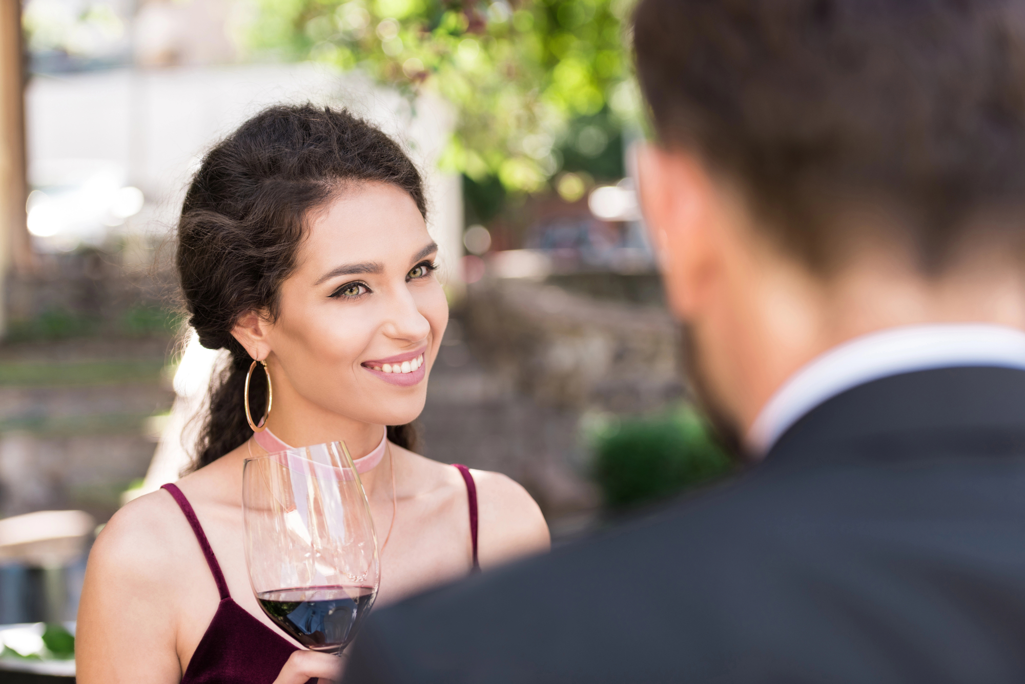 A woman with dark hair smiles while holding a glass of red wine, looking at a man with his back to the camera. The scene is outdoors with greenery in the background. The woman is wearing a sleeveless top and large hoop earrings.