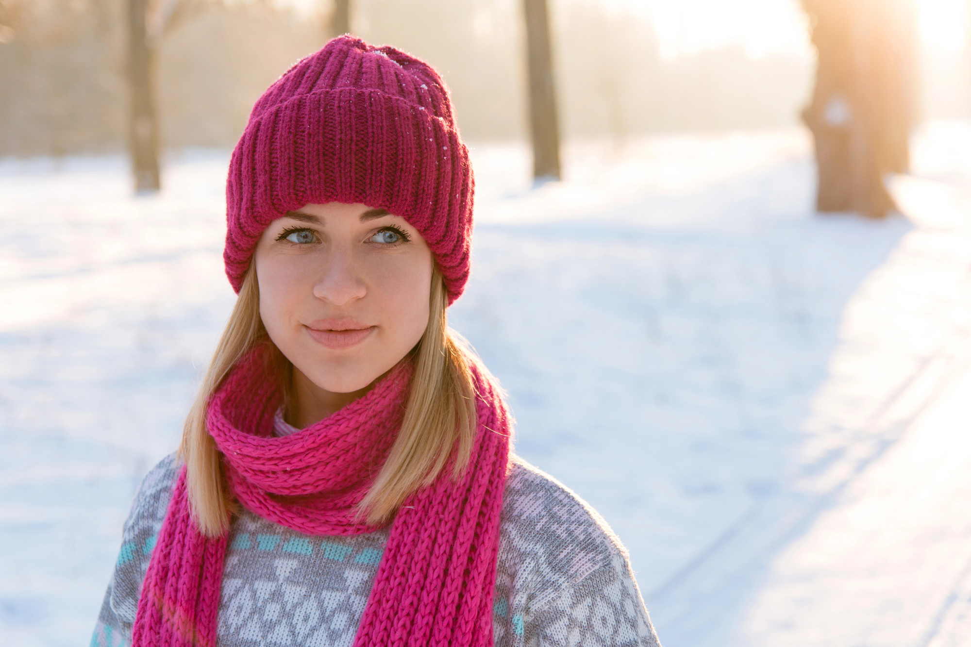 A young woman stands outdoors on a snowy day, wearing a pink knitted hat and matching scarf. She has blonde hair and is dressed in a geometrically patterned sweater. Sunlight filters through trees in the background, casting a warm glow.