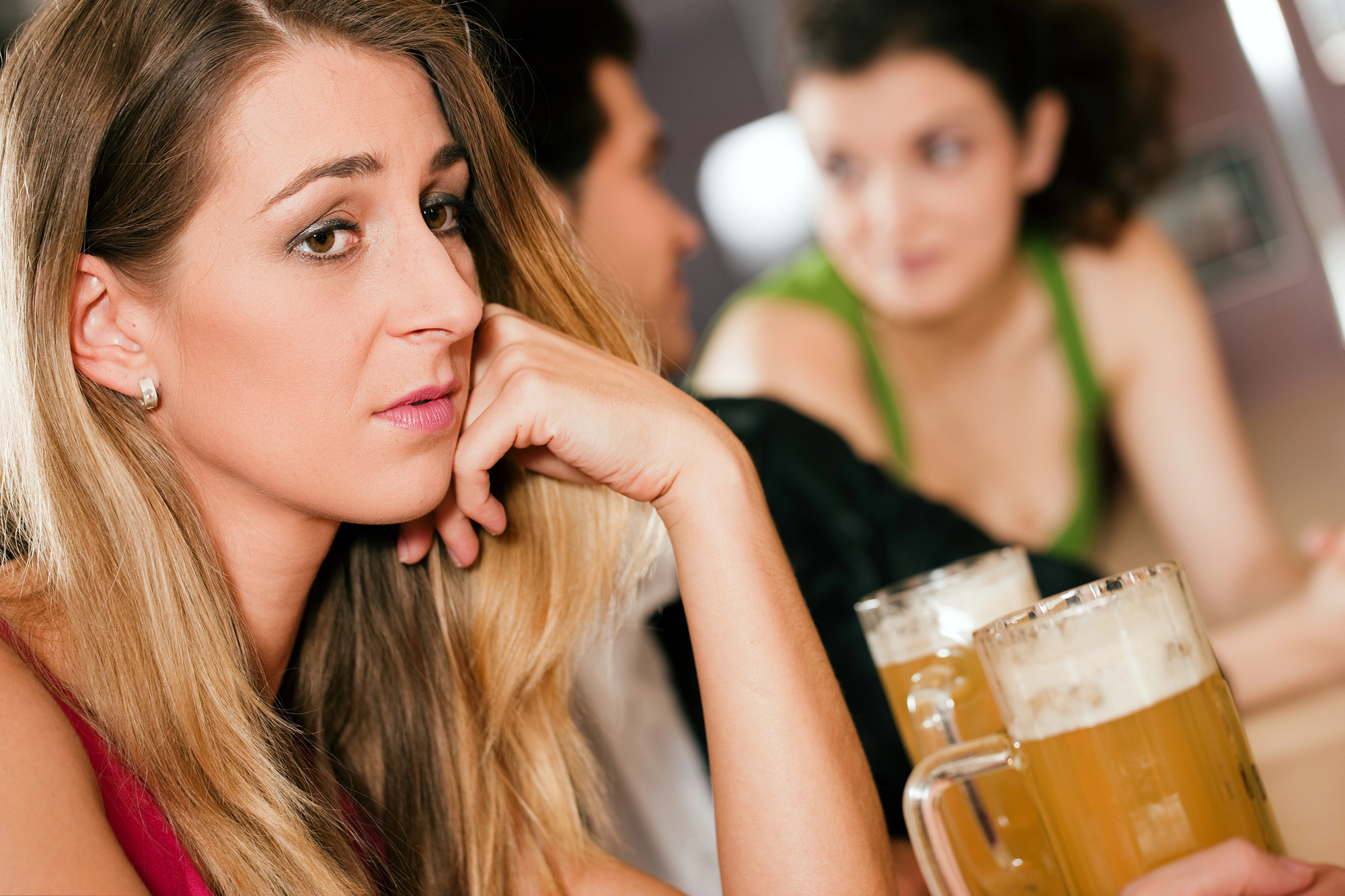 A woman with long blonde hair, dressed in a sleeveless top, looks thoughtful and concerned as she sits at a bar. In the background, a man and a woman with curly hair are having a conversation, and two glasses of beer are placed on the counter.