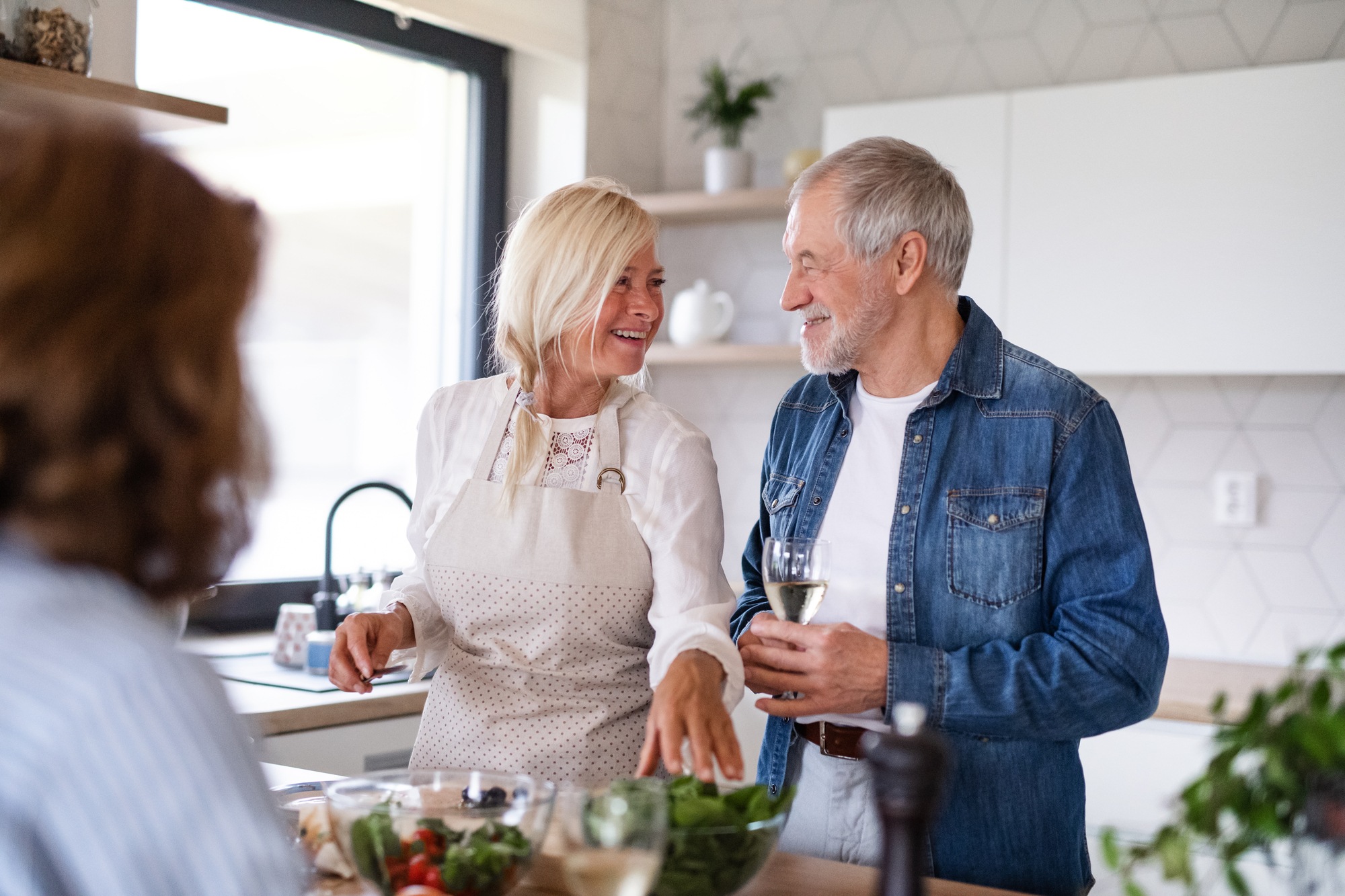 An older woman and man are enjoying time together in a modern kitchen. The woman, wearing an apron, gestures towards the counter filled with various vegetables. The man, dressed in a denim jacket, holds a glass of white wine. Another person is blurred in the foreground.