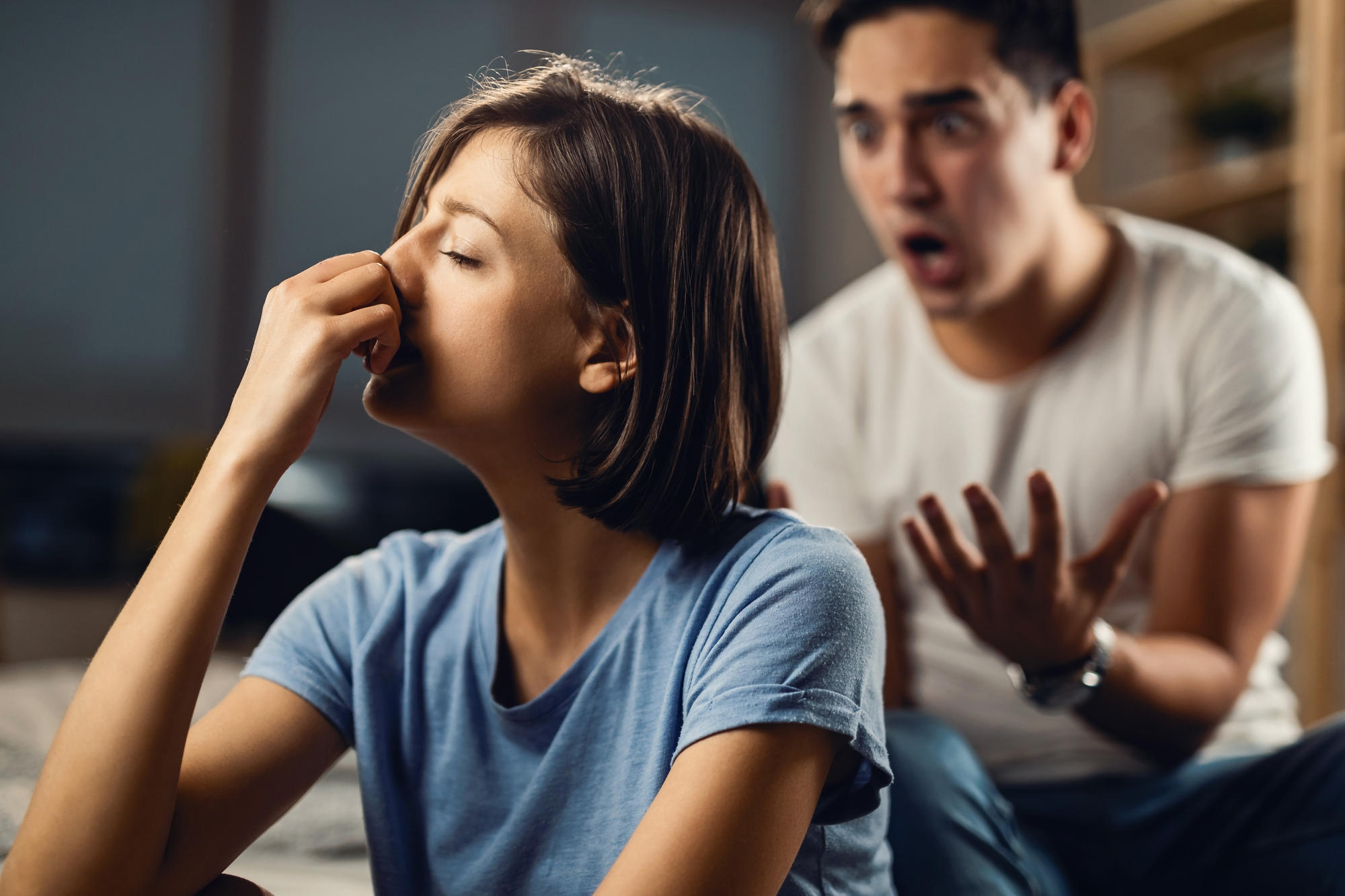 A woman with short brown hair in a blue shirt is pinching the bridge of her nose, appearing frustrated. In the background, a man with short dark hair in a white shirt has a distressed expression, with his arms raised as he seems to be arguing.