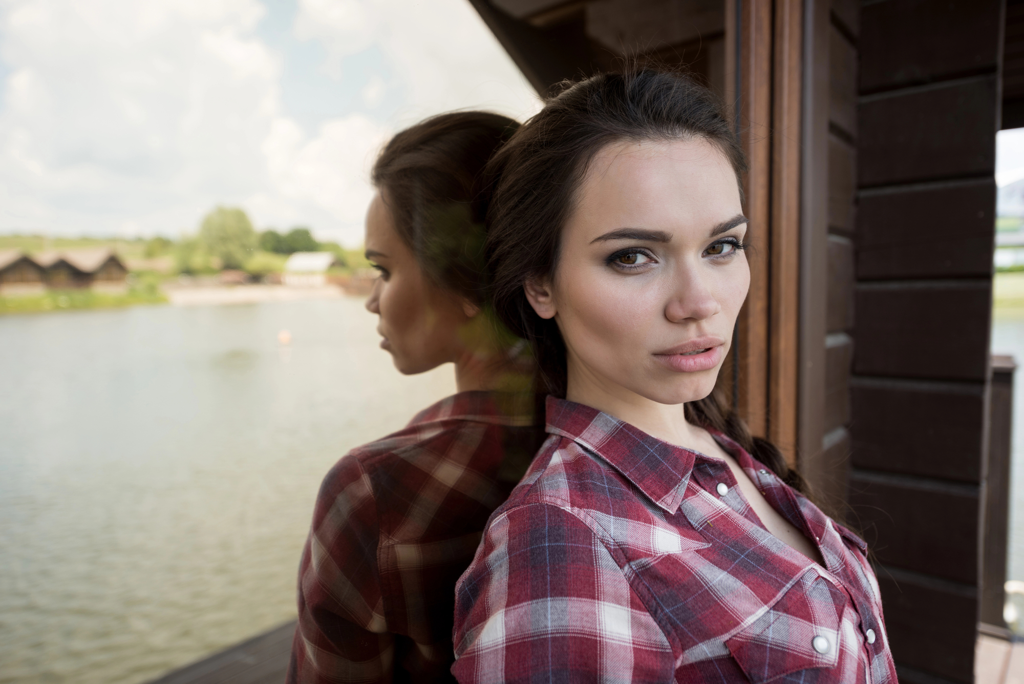 A woman in a red plaid shirt stands against a wooden structure near a calm lake. Her reflection is visible in the glass beside her. The background shows a few huts on the grassy bank and a partly cloudy sky. She has a serious expression on her face.