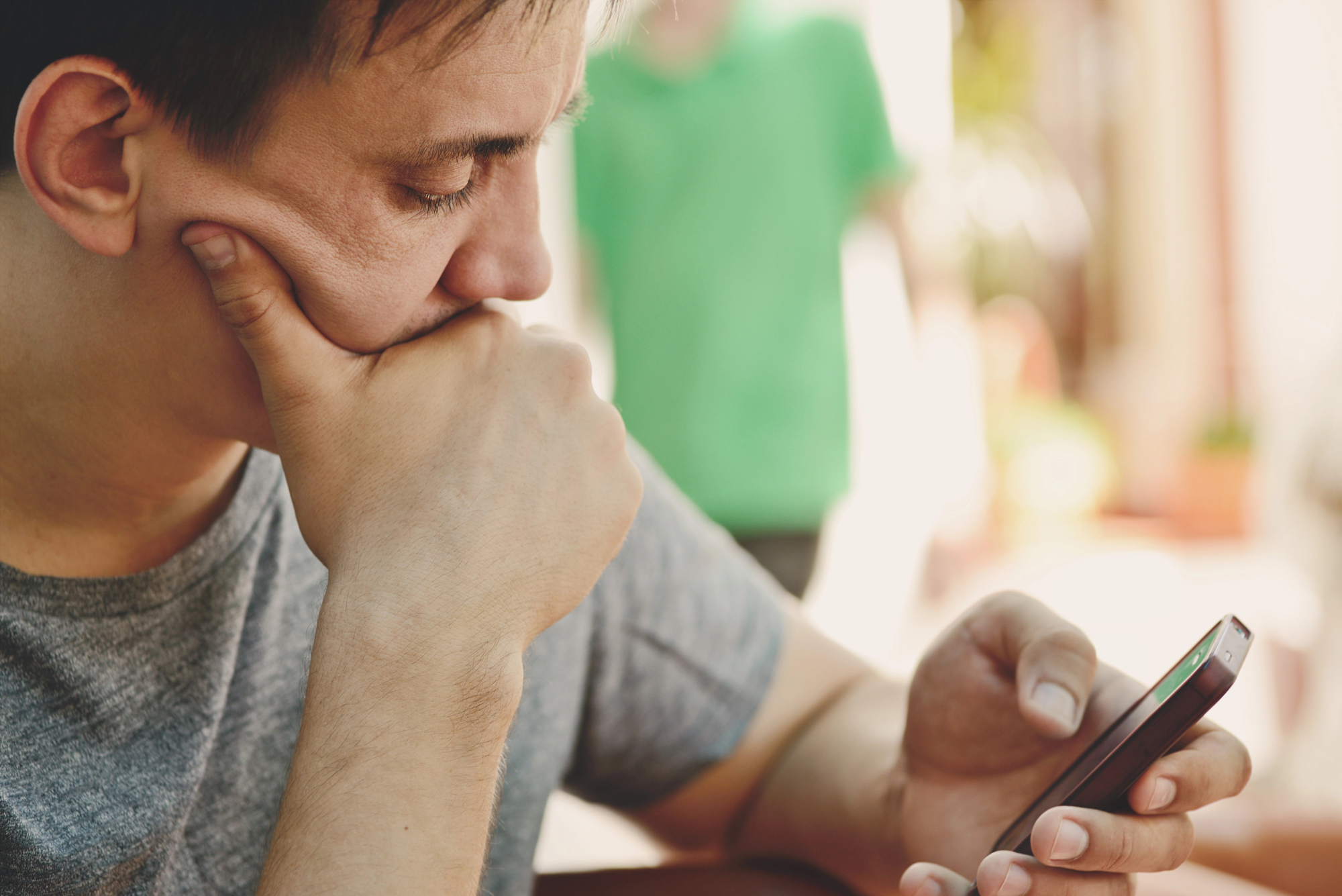 A person with short hair and a grey shirt is focusing intently on a smartphone. They rest their chin on their hand in contemplation. The background is blurred but shows an individual in a green shirt and an outdoor setting.