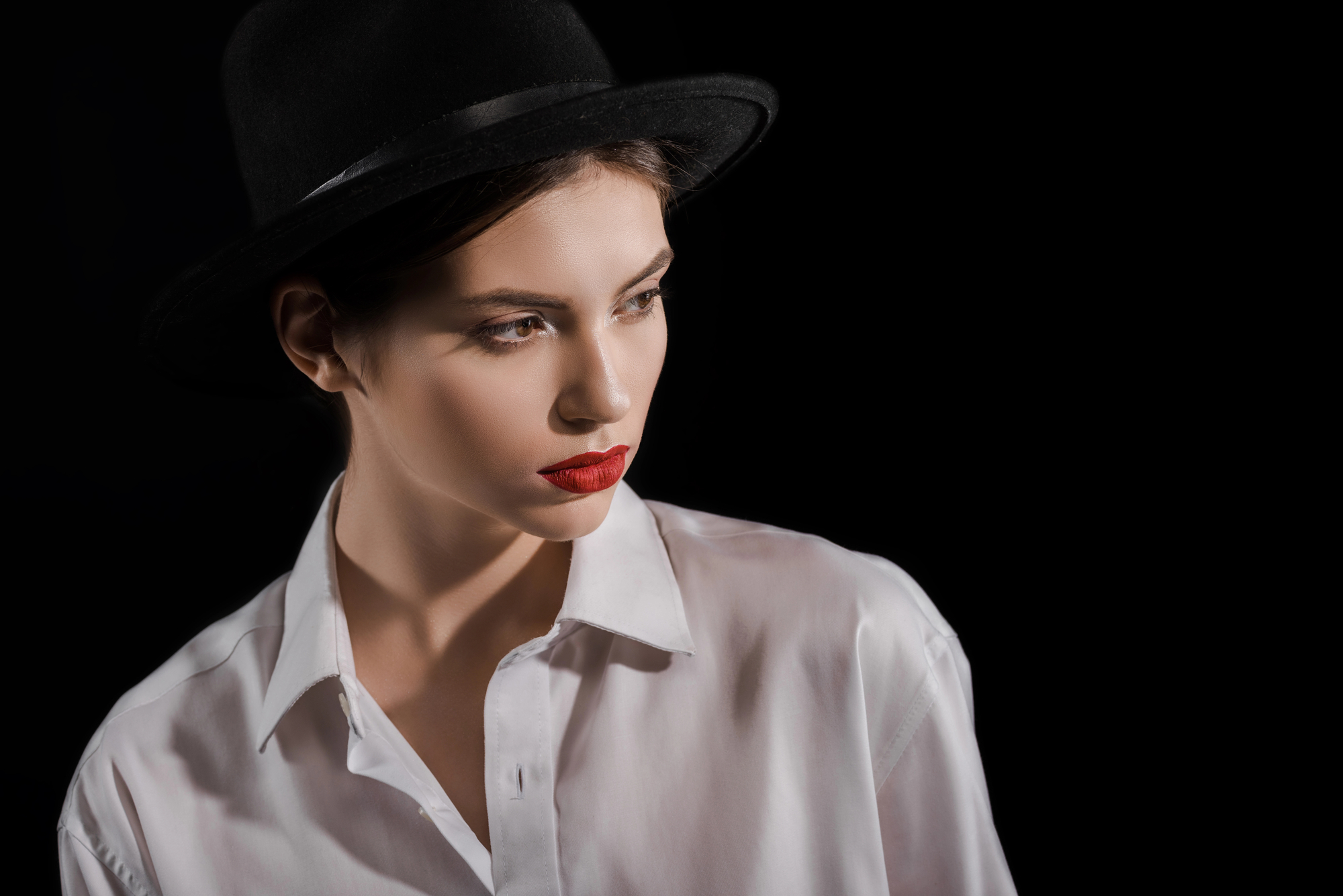 A woman wearing a black hat and white button-up shirt is looking off to the side against a dark background. She has red lipstick and a serious expression. The lighting highlights her facial features, casting soft shadows.