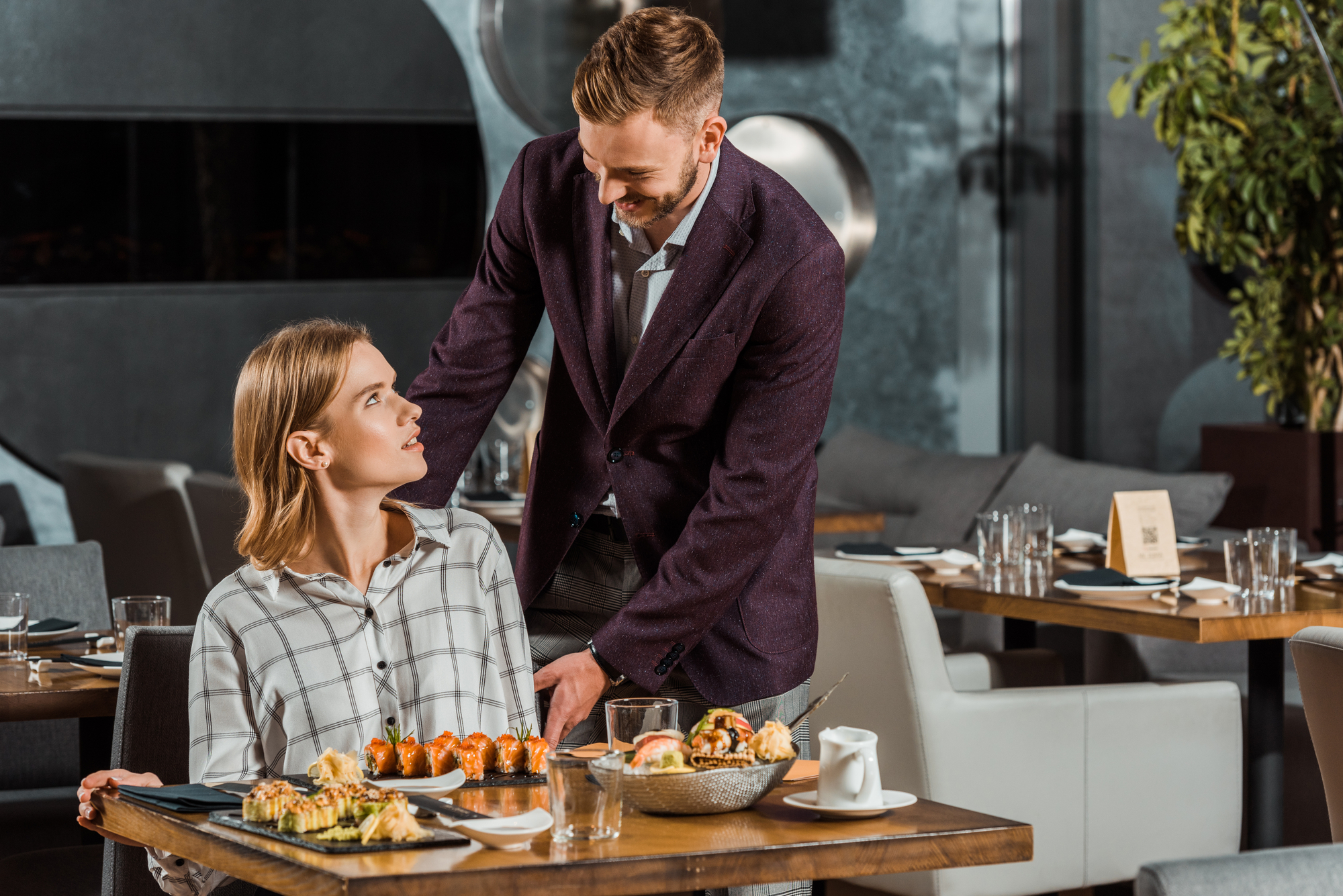 A woman and a man are in a modern restaurant. She is seated at a table with sushi and a cup of tea, looking up at him. The man, in a dark blazer, stands next to her, leaning forward slightly, engaged in conversation. The restaurant has a stylish, contemporary interior.