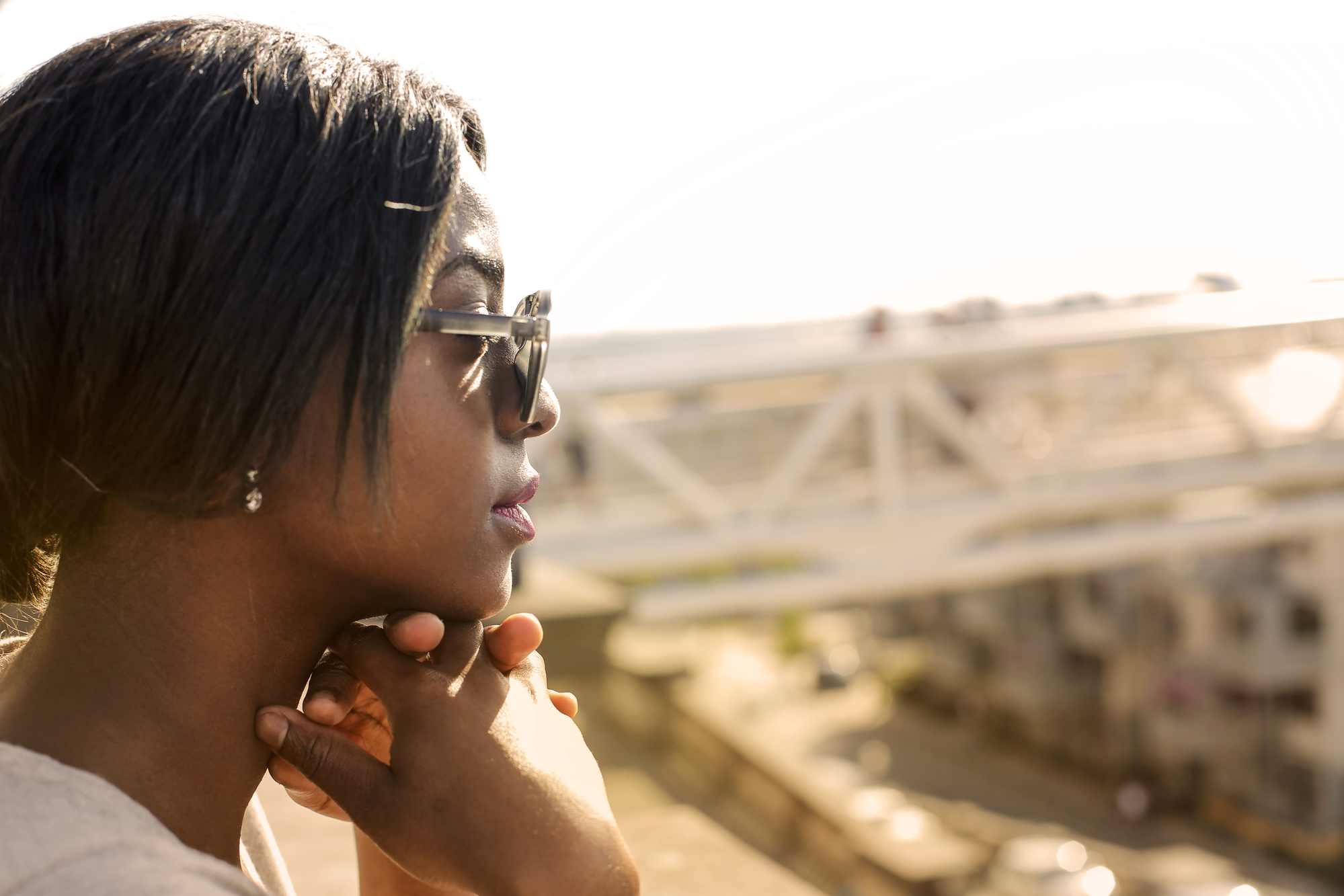 A person wearing sunglasses and a white top is shown in profile, resting their chin on their hands. They are outside, with blurred background elements of an industrial, urban setting featuring white bridge structures and buildings.