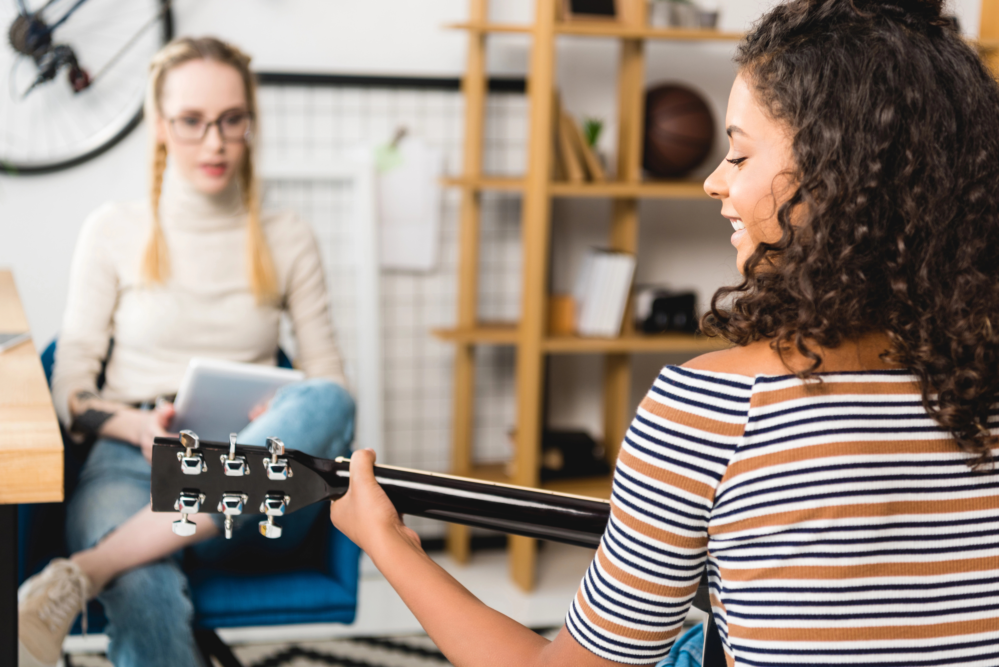 Two women are in a cozy room with bookshelves. One is playing an acoustic guitar, wearing a striped shirt, and has curly hair. The other, dressed in a white turtleneck and jeans, is sitting and holding a tablet, watching her.