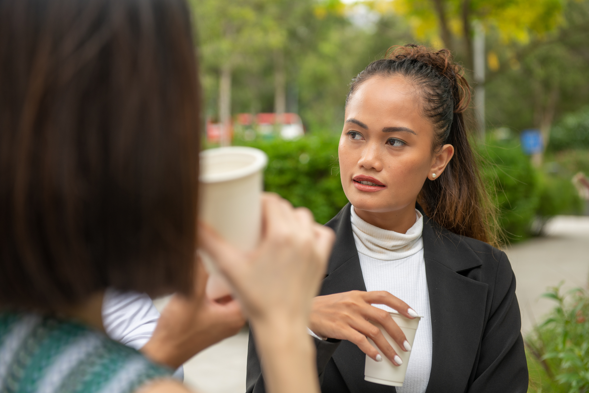 A woman with dark hair tied back is holding a cup and listening attentively to another person in the foreground who is also holding a cup. They appear to be having a conversation outdoors with green foliage and trees in the background.