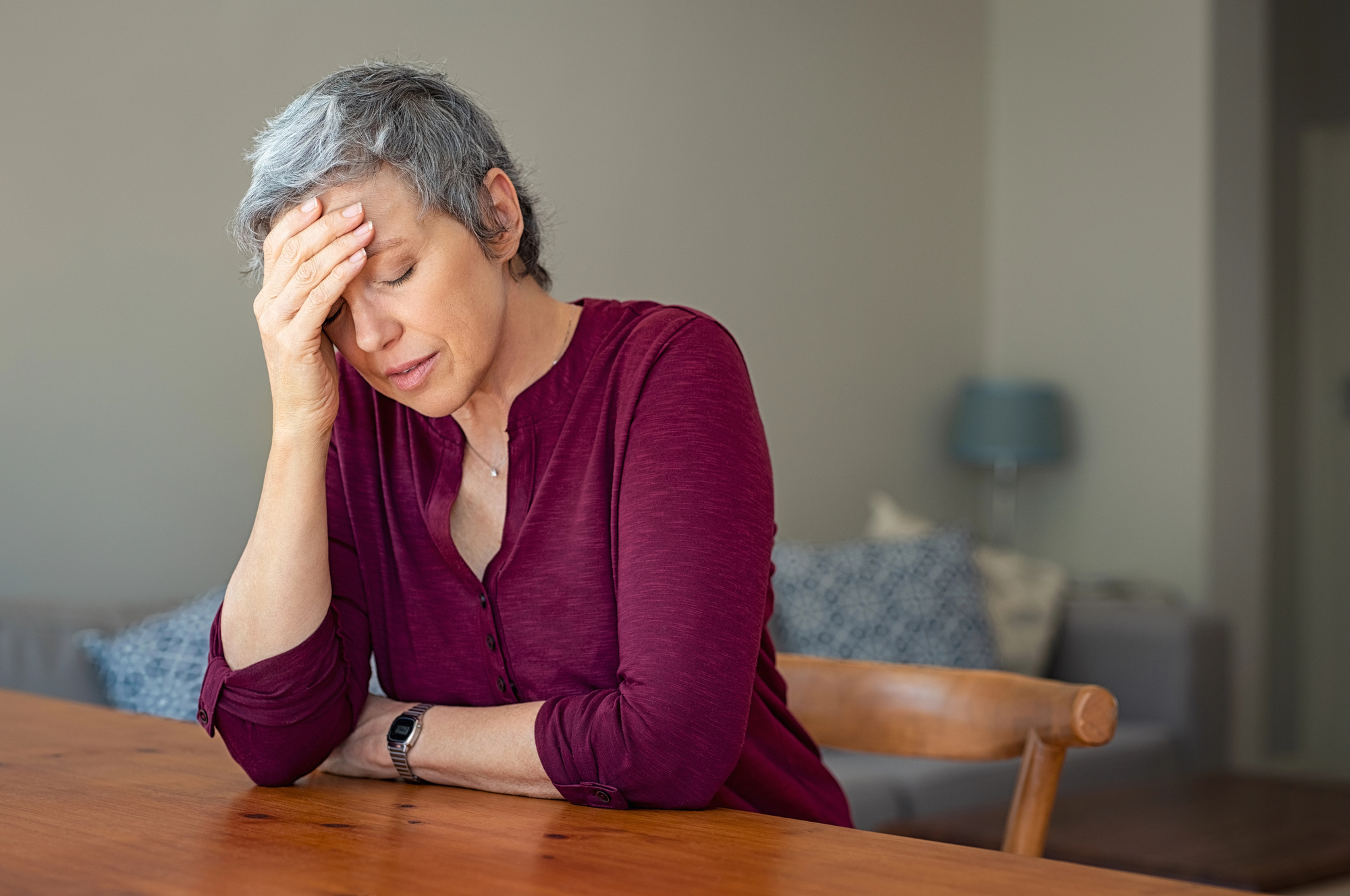 A woman with short gray hair and wearing a maroon shirt sits at a wooden table with her hand on her forehead, eyes closed, appearing stressed or tired. The background features a blurred living room with a lamp and cushions.