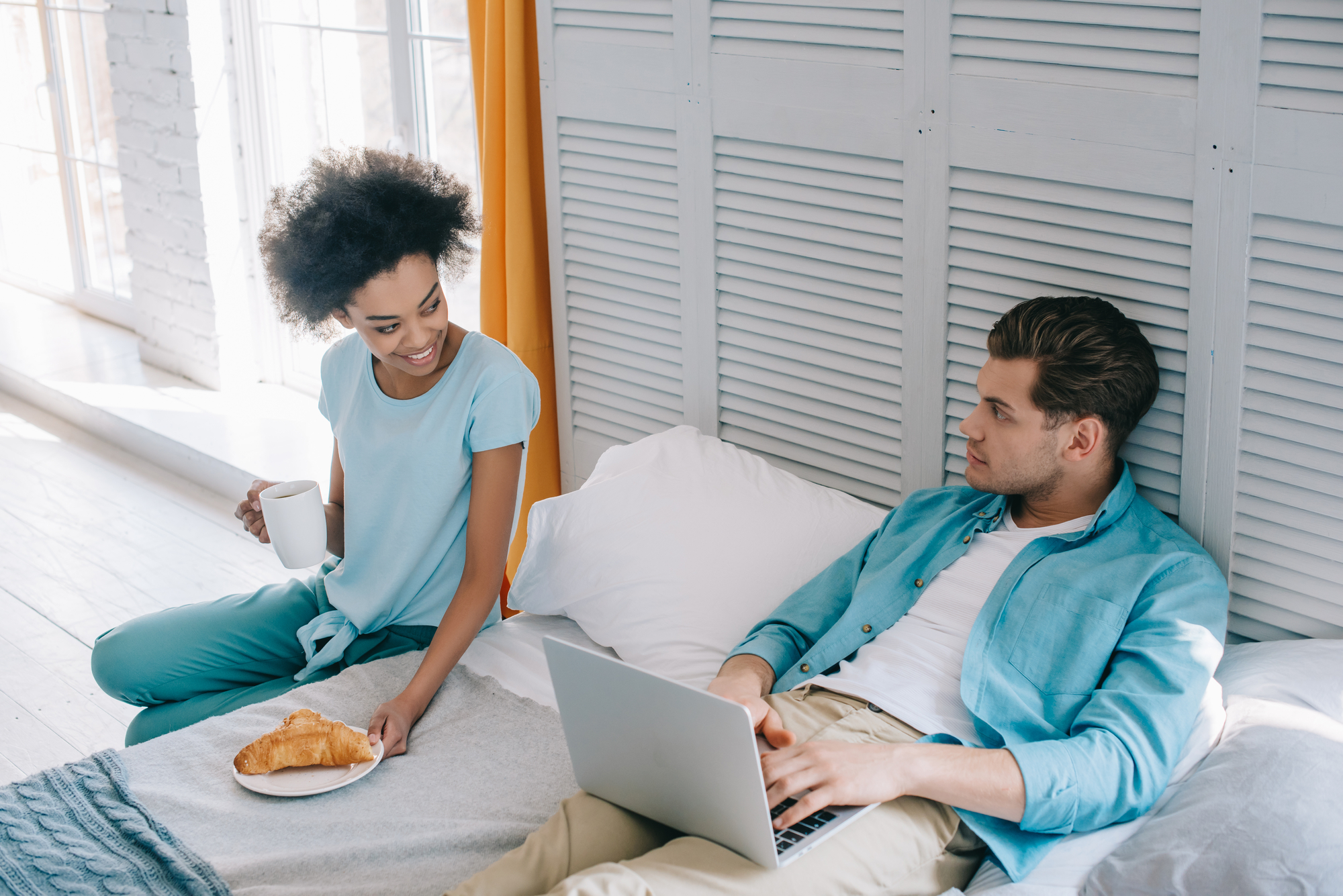 A woman holding a cup and a plate with a croissant stands next to a bed while smiling at a man who is lying on the bed, using a laptop. The room is bright with natural light from a large window, and there are white walls and yellow curtains.