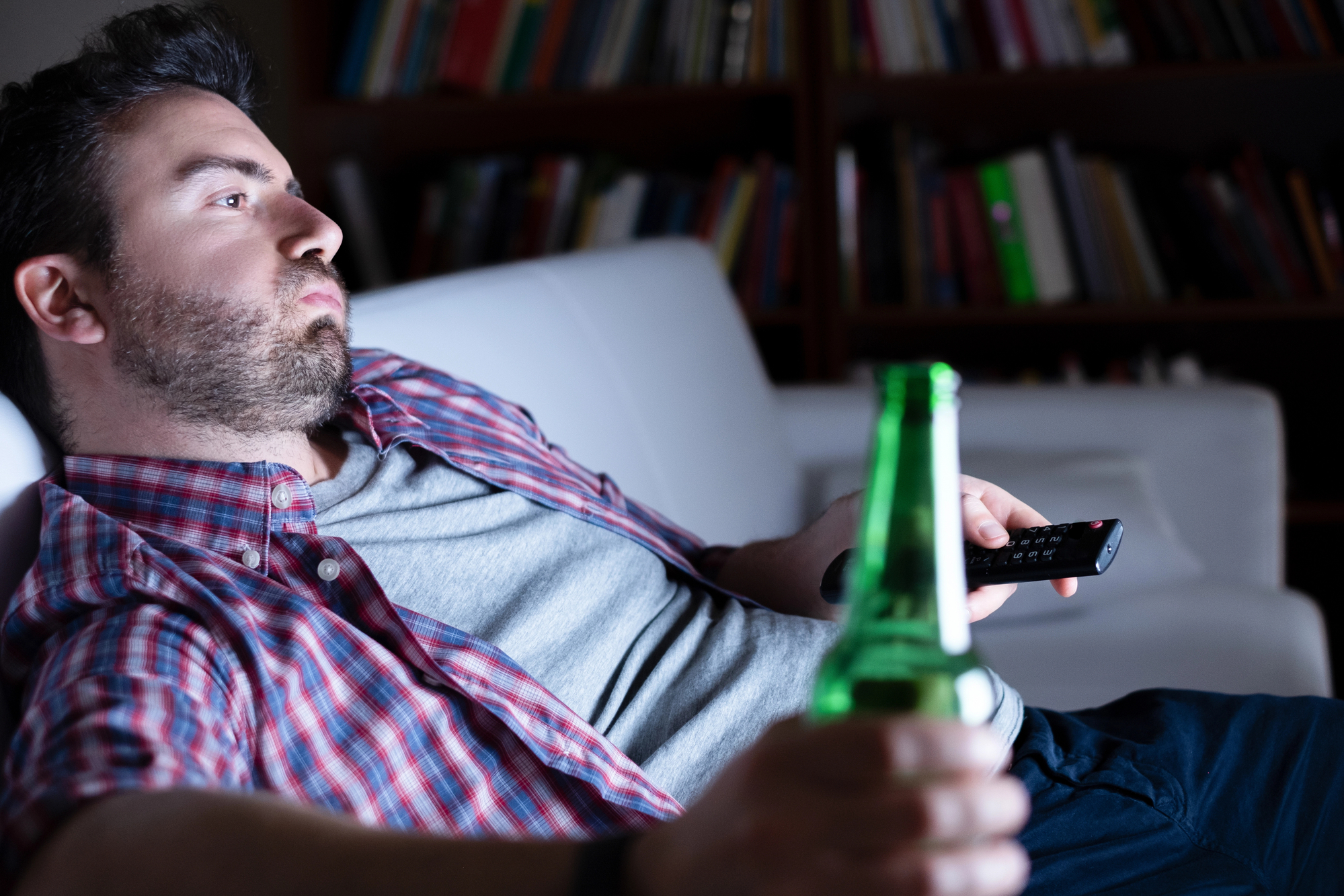 A man with a beard lounges on a white couch, holding a remote control in one hand and a green beer bottle in the other. He is wearing a red plaid shirt over a grey t-shirt. A bookshelf filled with books is blurred in the background.