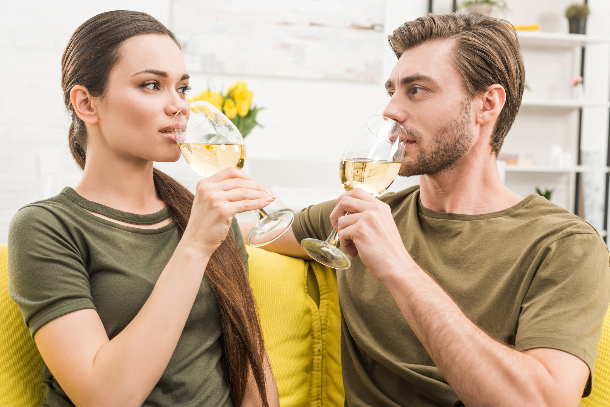 A woman and a man are sitting on a yellow couch, both wearing green shirts, and enjoying glasses of white wine. They are facing each other, smiling, and appear to be in a relaxed and comfortable setting with a bright, modern background.