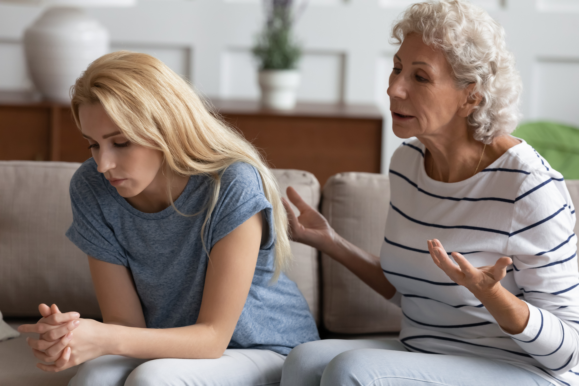 A senior woman with gray curly hair passionately talks to a young blonde woman sitting next to her on a couch. The young woman looks down, appearing upset or deep in thought. The room has subtle decor with a potted plant and a white vase in the background.