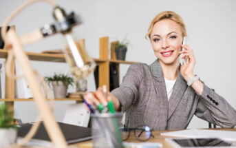 A woman in a plaid blazer smiles while talking on the phone at her desk. She reaches for a pen from a holder. The desk has office supplies, a pair of glasses, and a decorative lamp. Shelves with plants and folders are visible in the background.