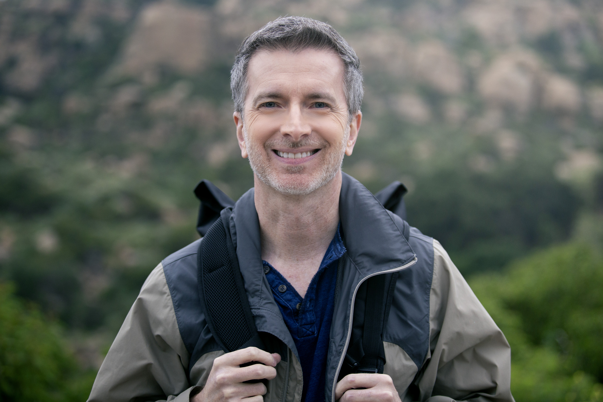 A man with short gray hair is standing outdoors, smiling at the camera. He is wearing a gray and black jacket and has a backpack on. The background features blurry, green foliage and rocky terrain.