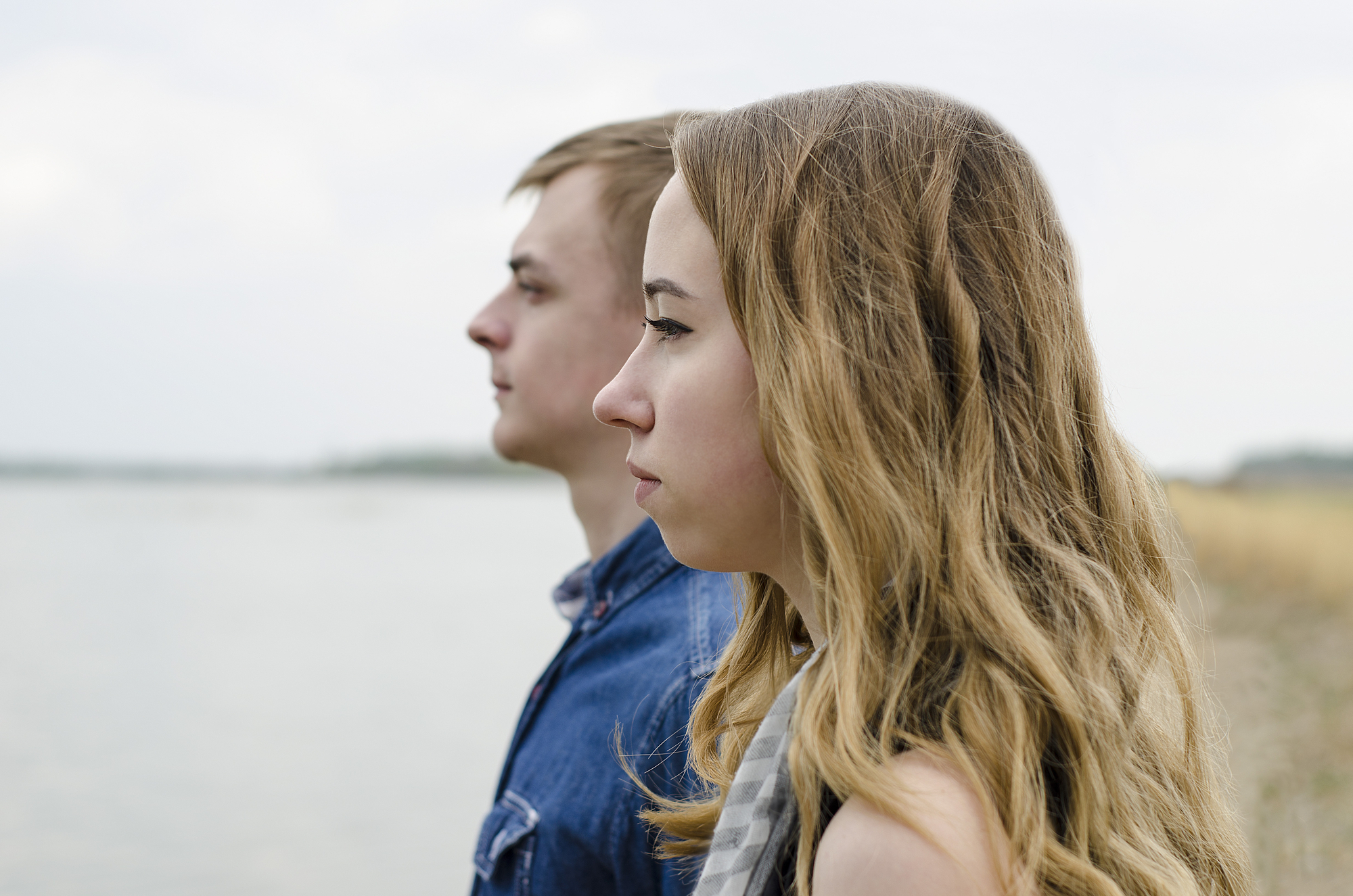 Two people with long hair, one in a blue shirt and the other in a black top, are standing side by side near a body of water. Both are gazing into the distance with a calm expression. The background is slightly blurred, showing the shoreline and a cloudy sky.